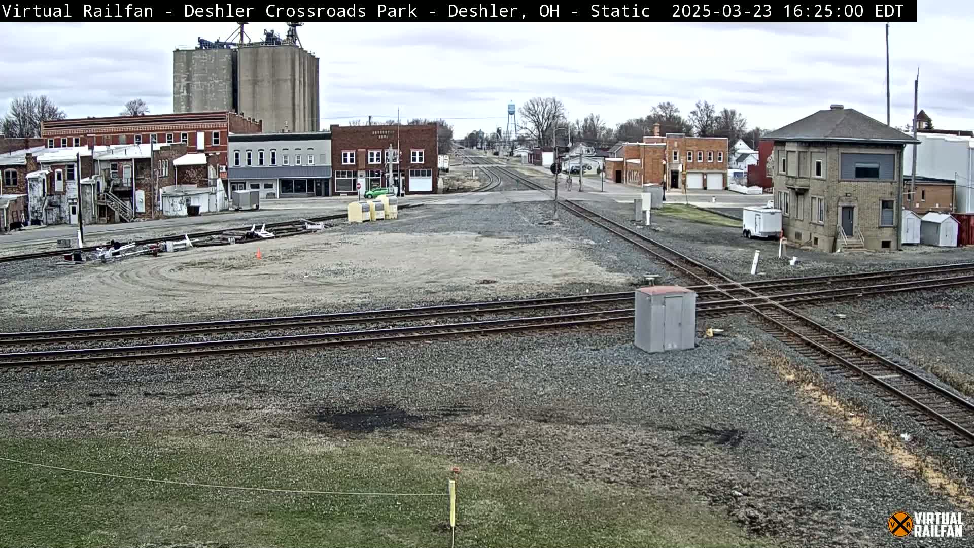 A view of a railway crossing in a small town on an overcast day, with several buildings and grain silos visible in the background.