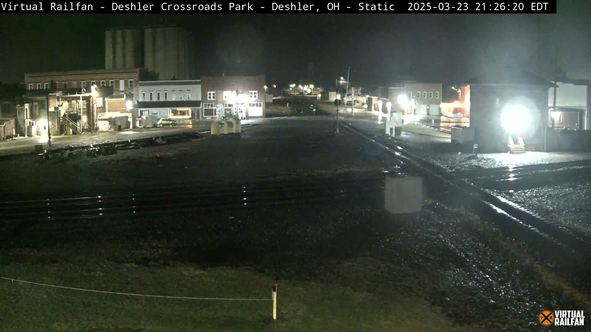 A nighttime view of a wet train crossing in a small town, with several buildings visible under dim lighting.