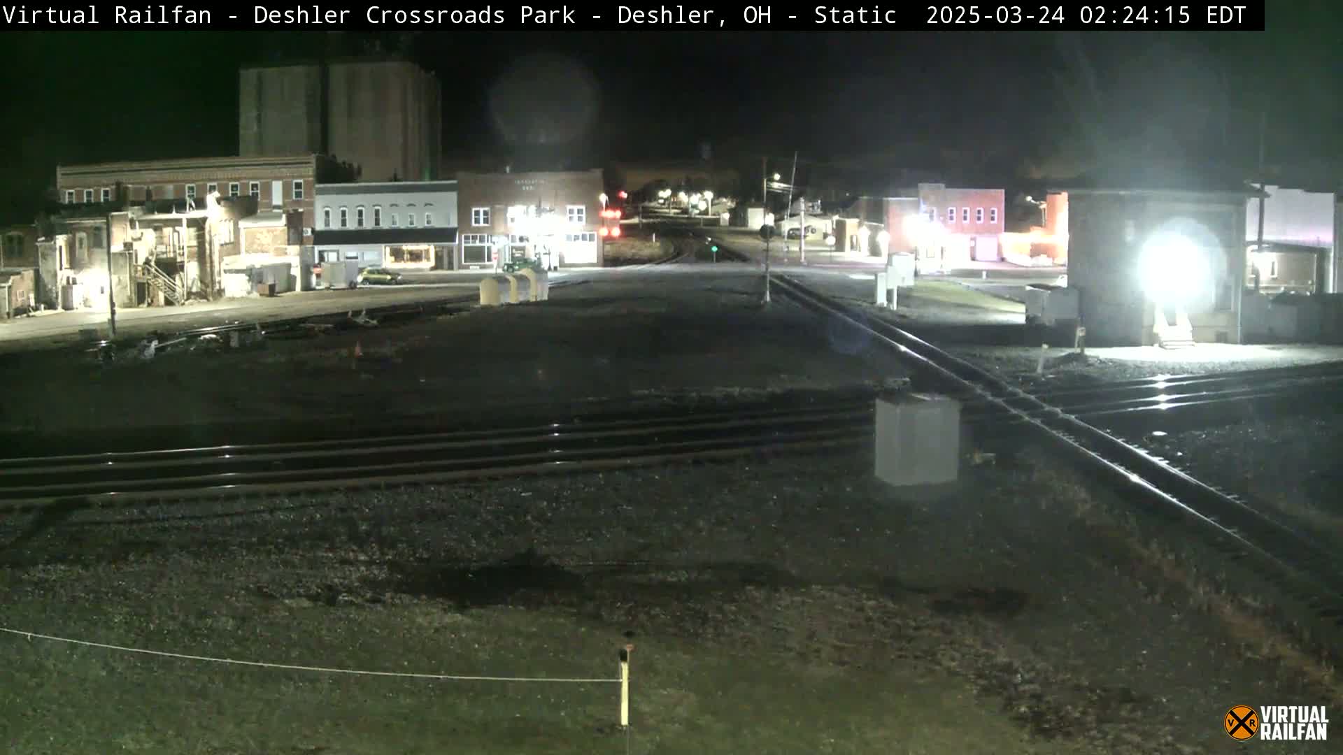 A nighttime view of a railroad crossing in a small town, with several buildings visible under dark skies.