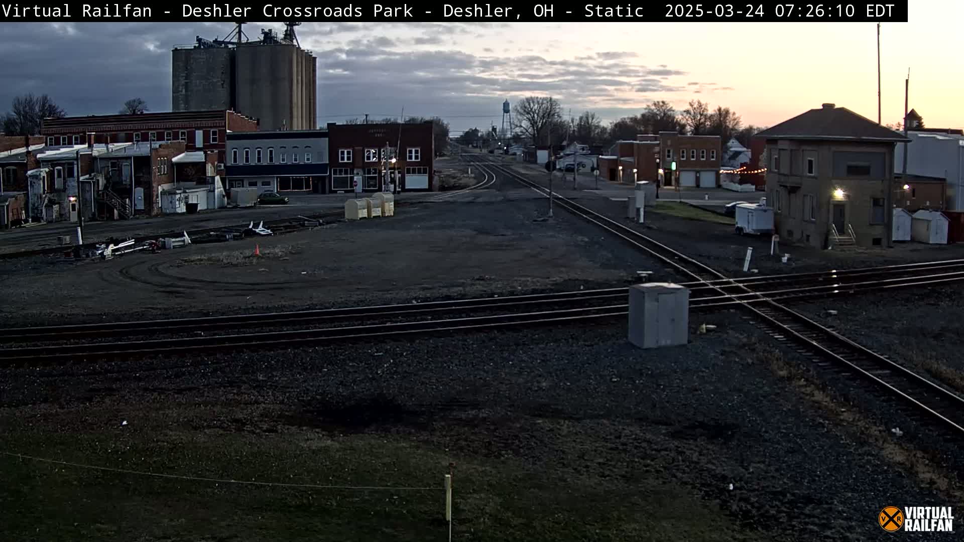 A railroad crossing in a small town at dawn, with several buildings, grain silos, and a railway station visible under a mostly cloudy sky.