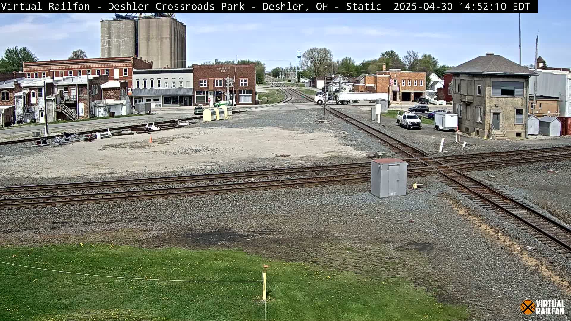 A sunny view of a railroad crossing in a small town, with several buildings, grain silos, and vehicles visible.