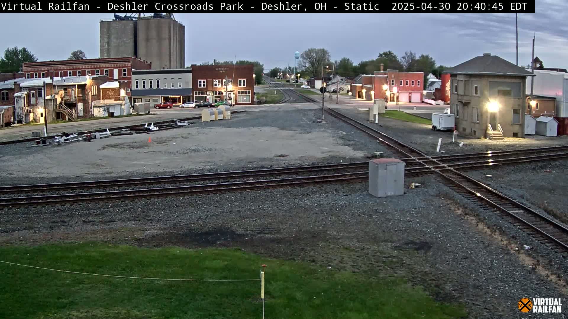 A twilight view of a railroad crossing in a small town, with several buildings visible and a grassy area in the foreground.