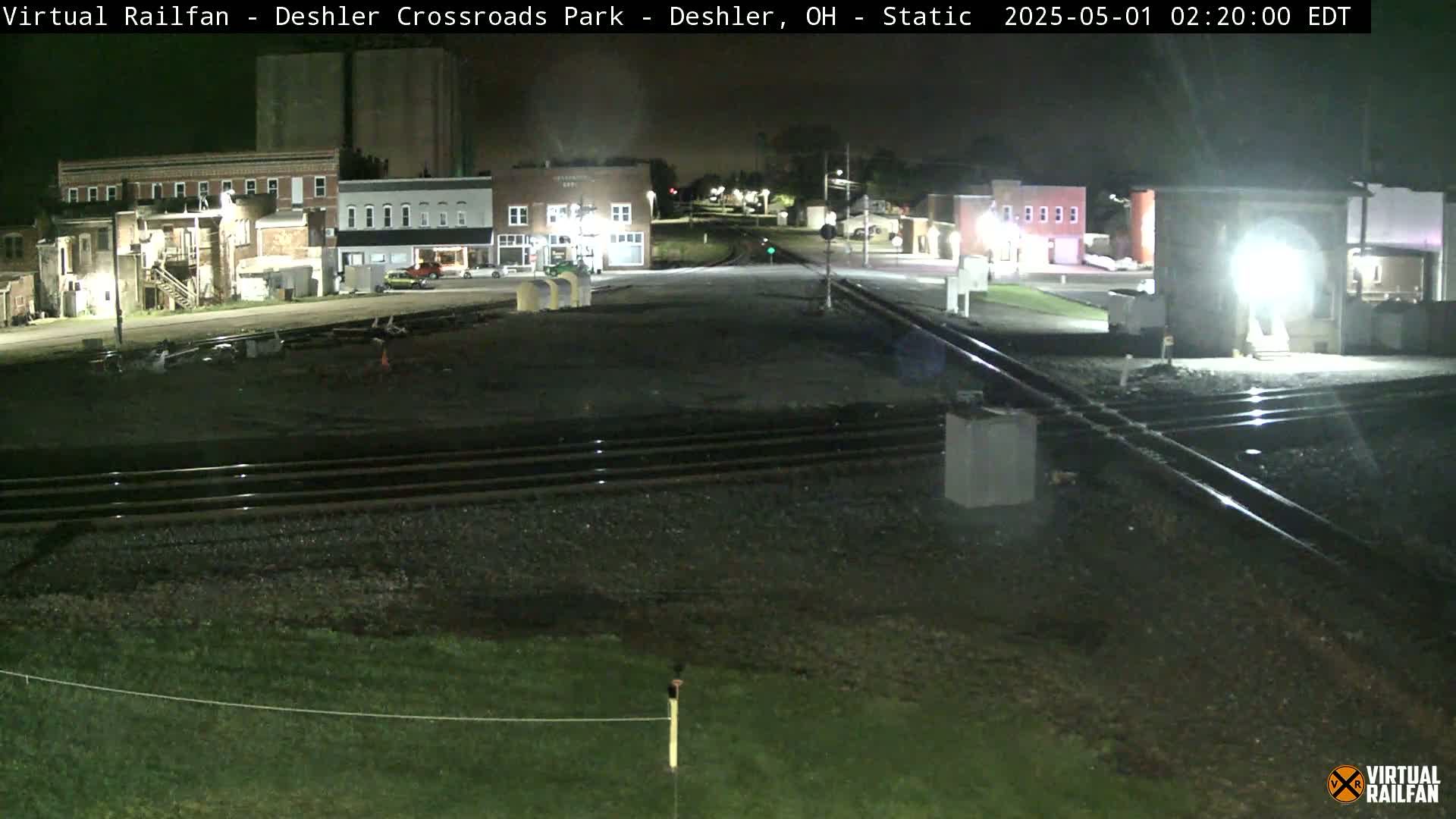A nighttime view of a railroad crossing in a small town, with several buildings visible under a dark sky.