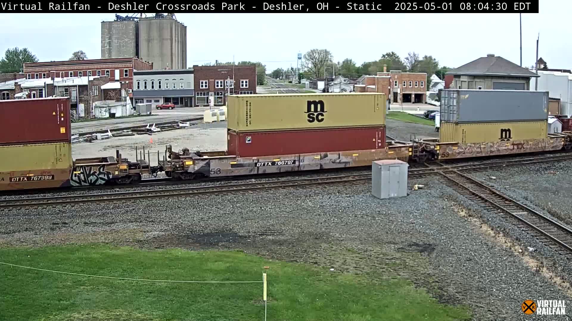 A freight train carrying shipping containers sits on railroad tracks in front of a small town under a mostly cloudy sky.