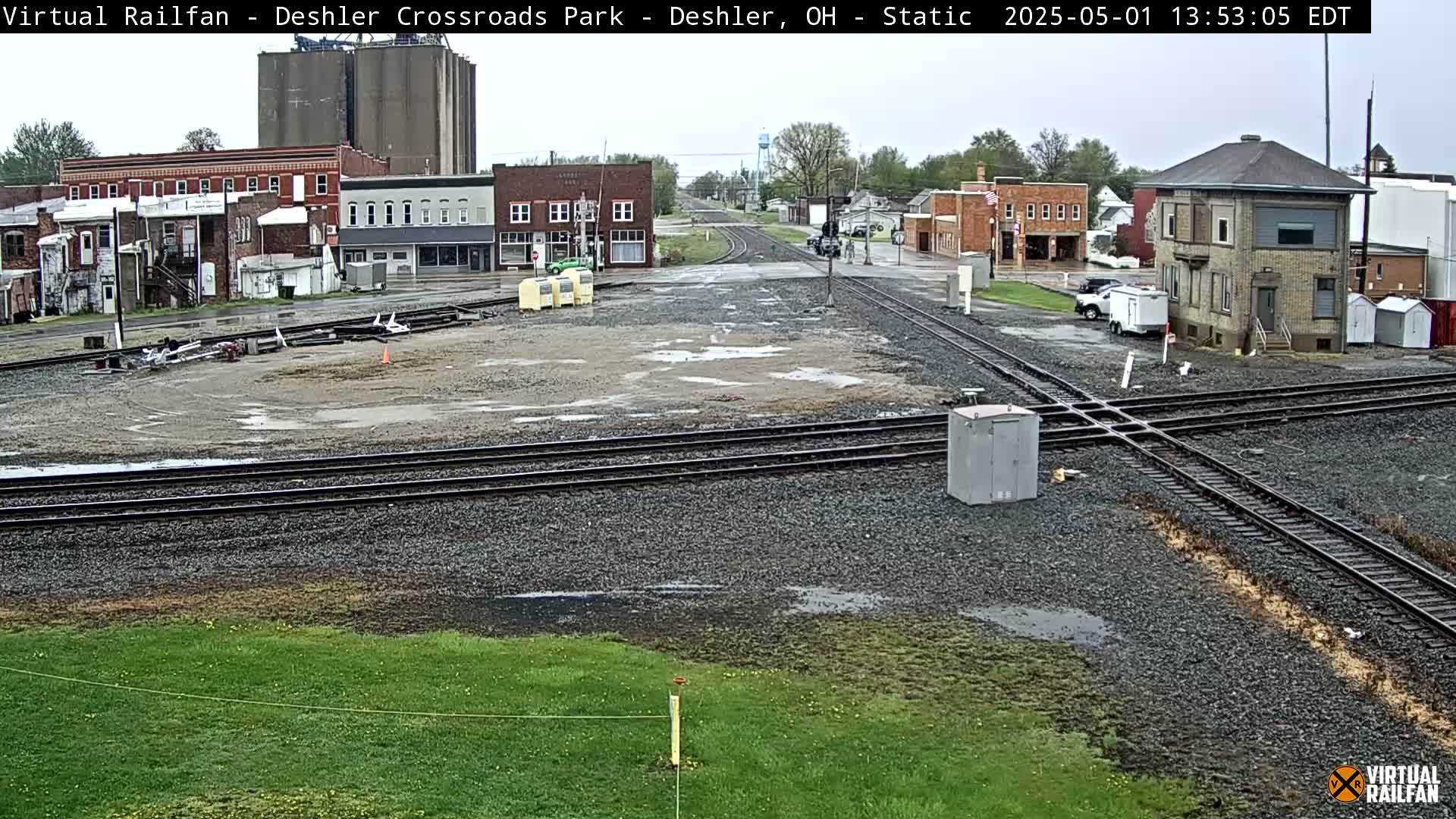 A wet, overcast day reveals a railroad crossing in a small town, with several buildings and grain silos in the background.