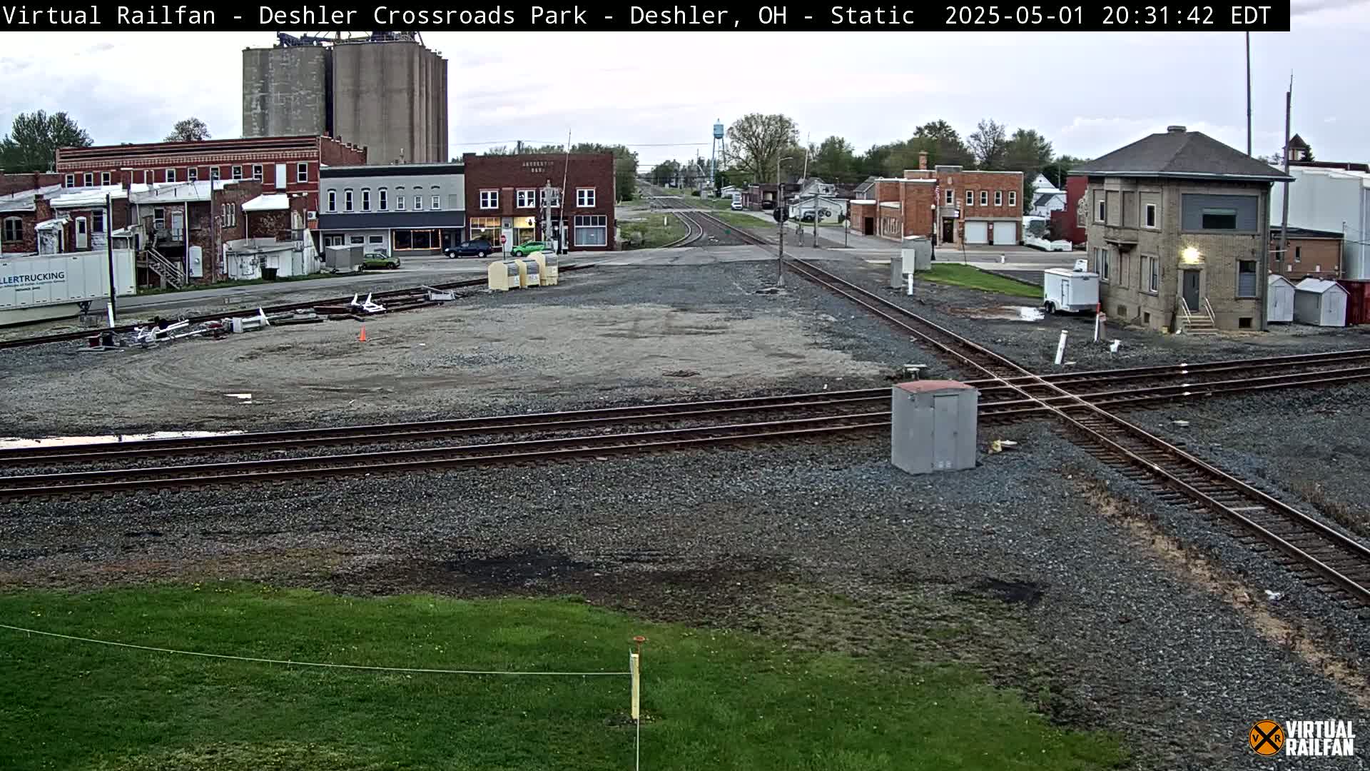 A railroad crossing in a small town under an overcast sky, with several buildings and grain silos visible in the background.