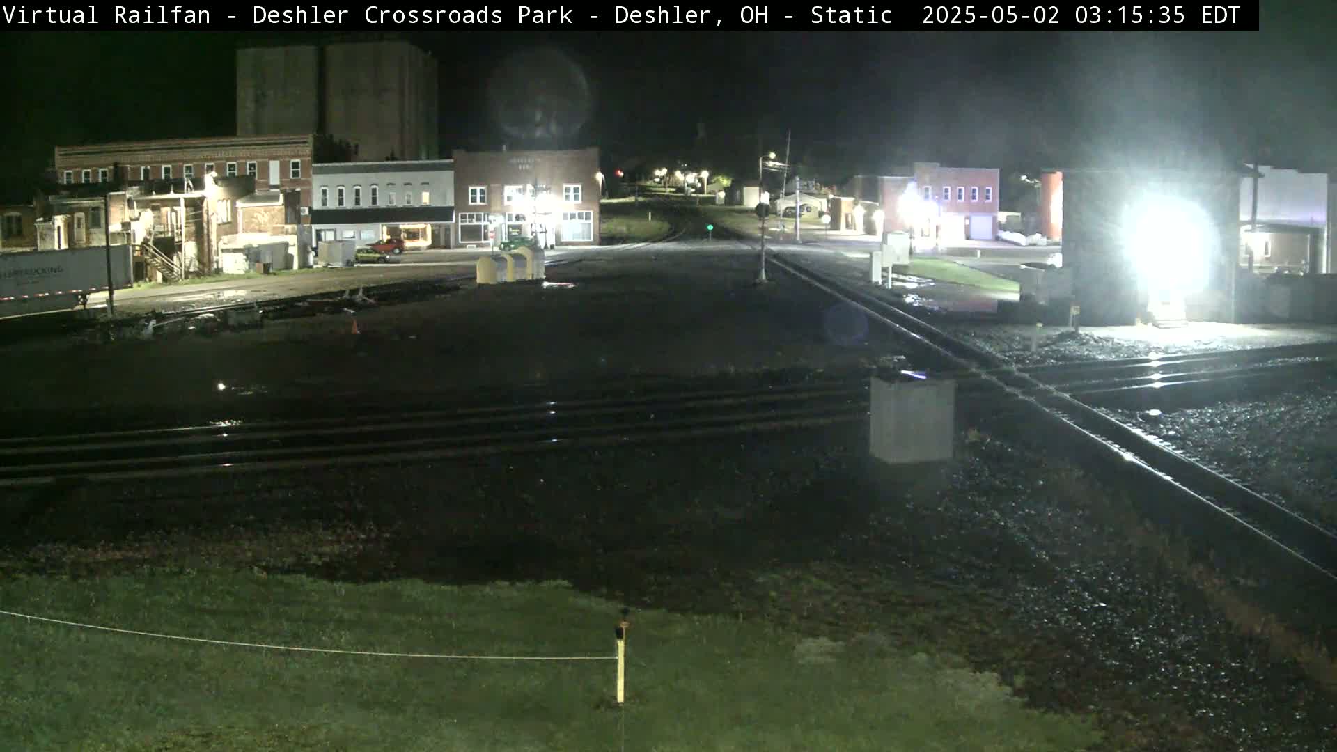 A nighttime view of a railroad crossing in a small town, with wet surfaces indicating recent rain.