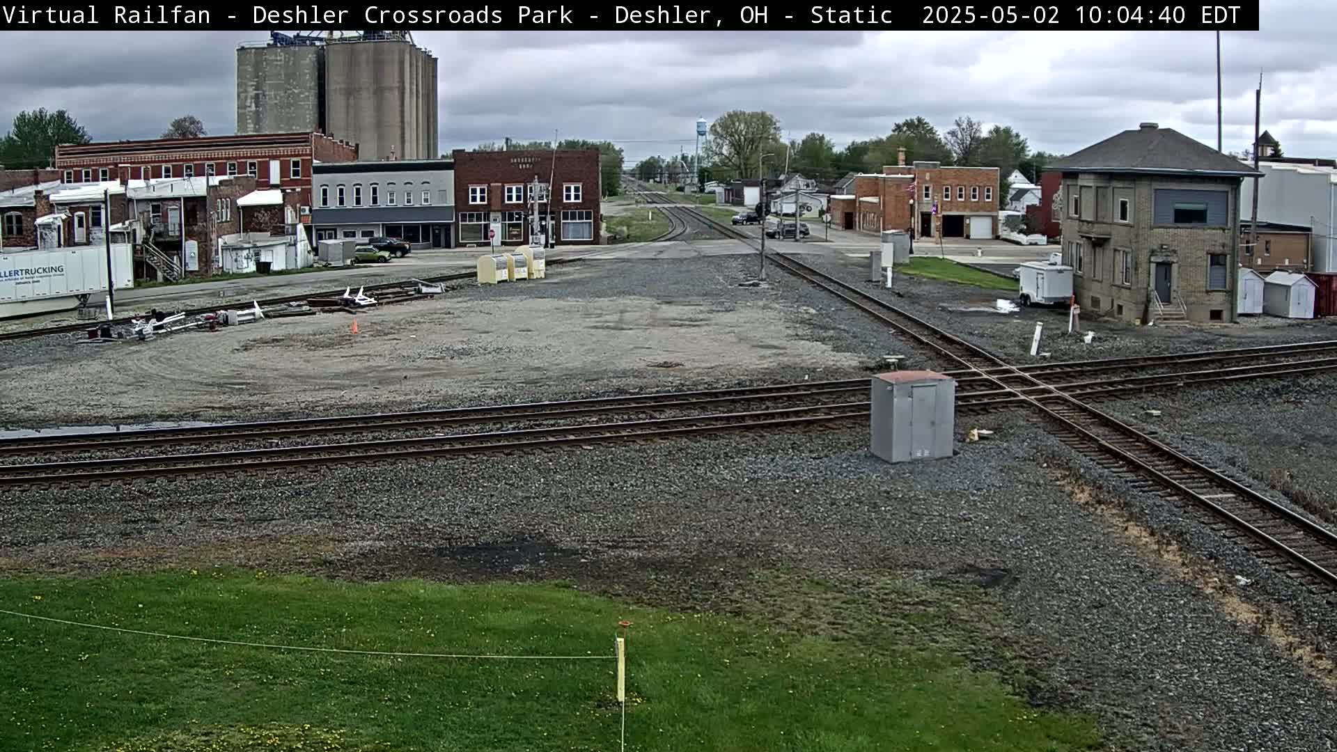 A railroad crossing in a small town under partly cloudy skies, with several buildings and grain silos visible in the background.