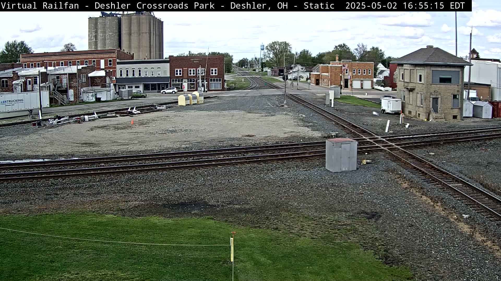 A railroad crossing in a small town on a partly cloudy day, with several buildings and grain silos in the background.