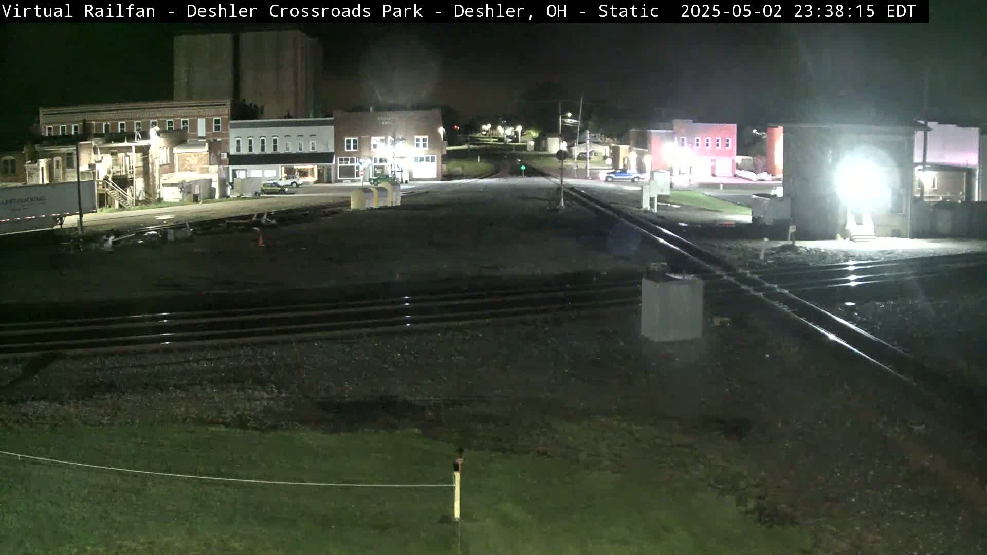 At night, a nighttime view shows a railroad crossing in front of several buildings under artificial light.