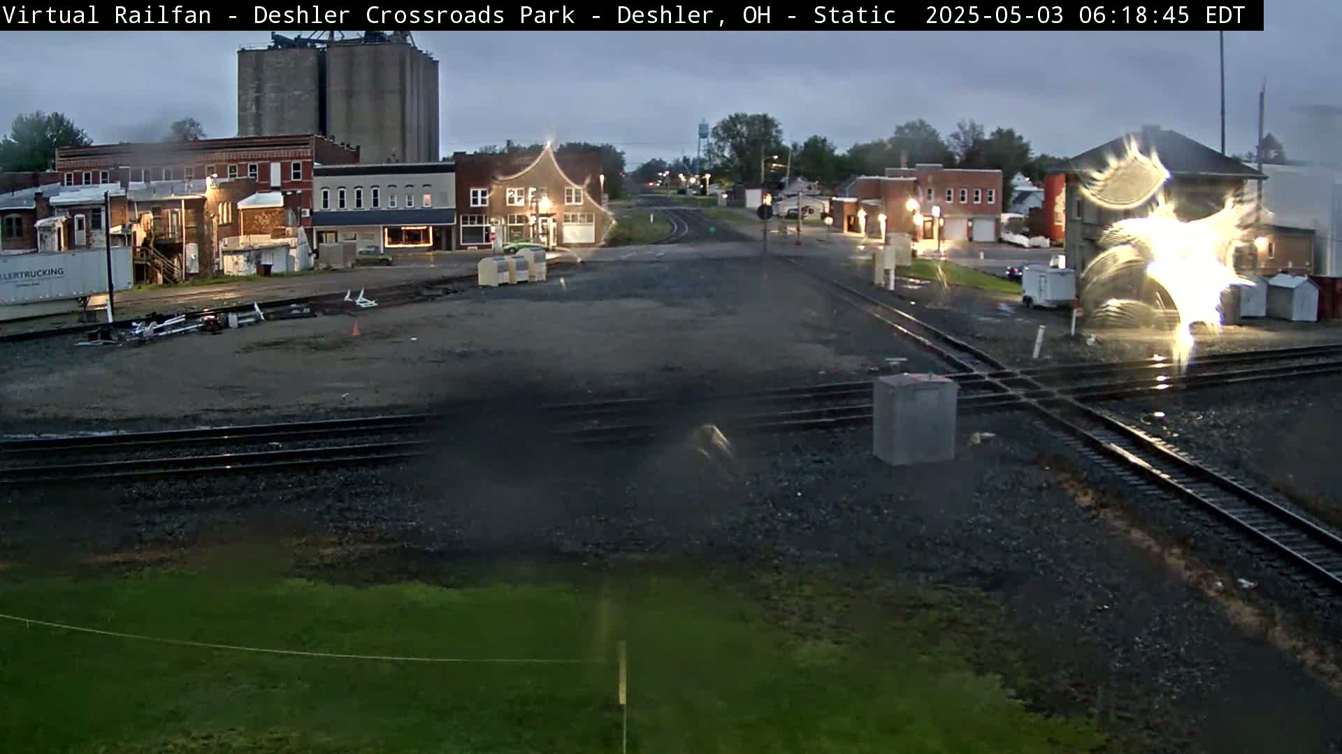 A railroad crossing in a small town on an overcast morning, with several buildings and grain silos visible in the background.