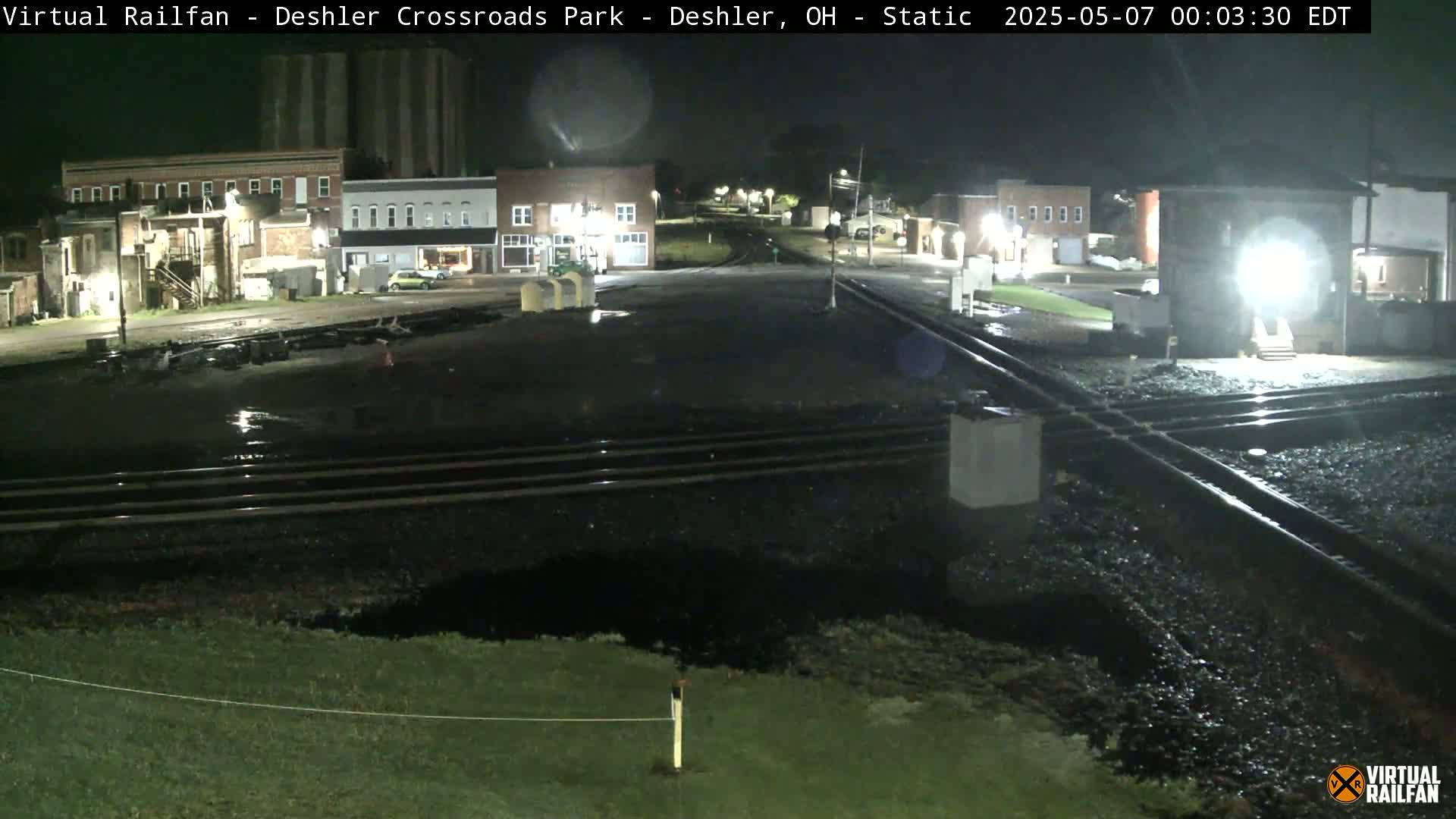 A nighttime view of a wet railroad crossing in front of several buildings, possibly in a small town.