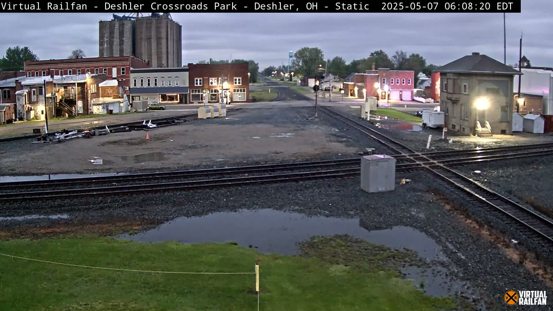 A wet, overcast morning scene shows a railroad crossing in a small town with several buildings, grain silos, and puddles of water.