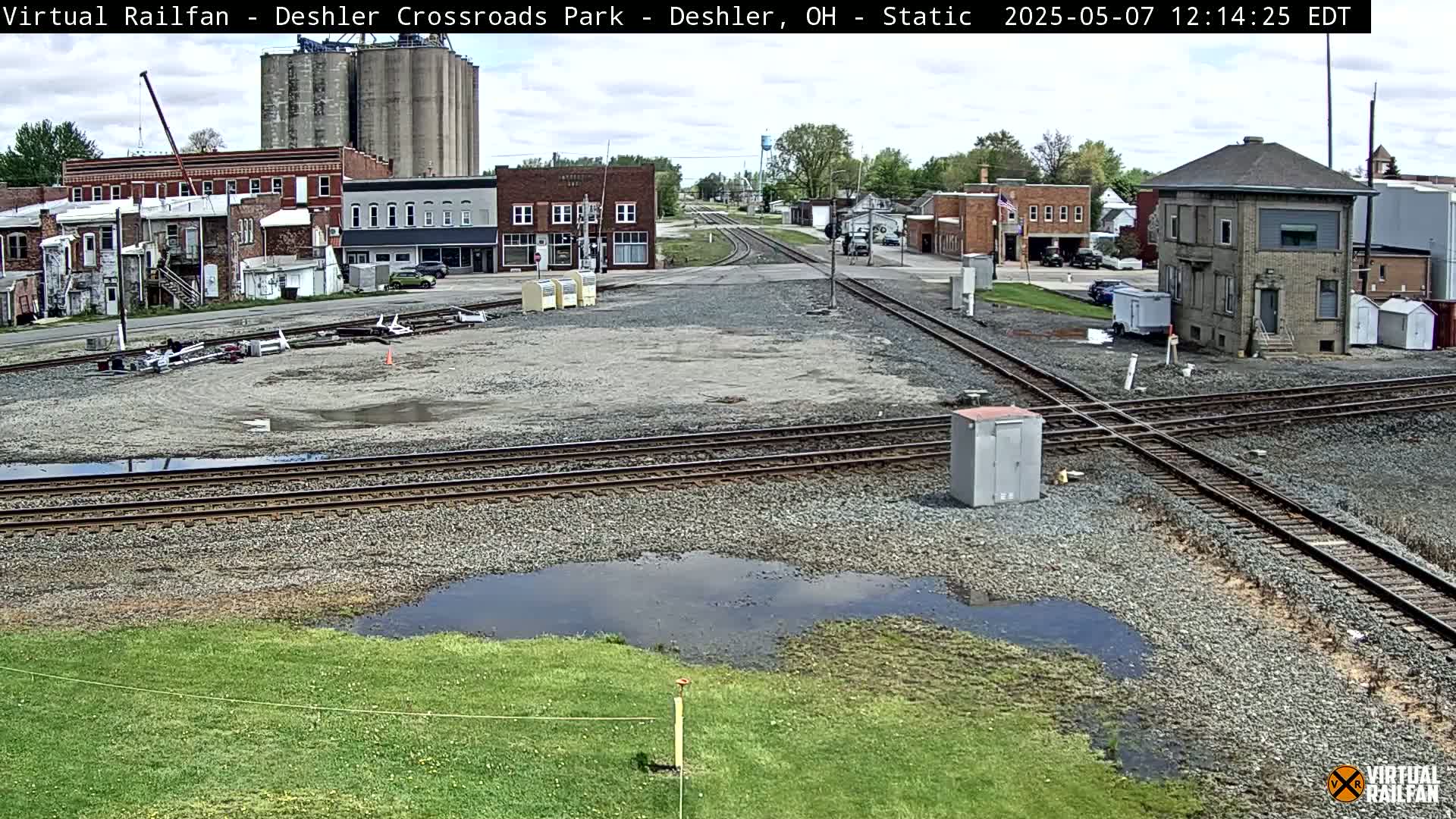 A mostly clear day shows a train crossing in a small town with several puddles of water on the ground near some railroad tracks.