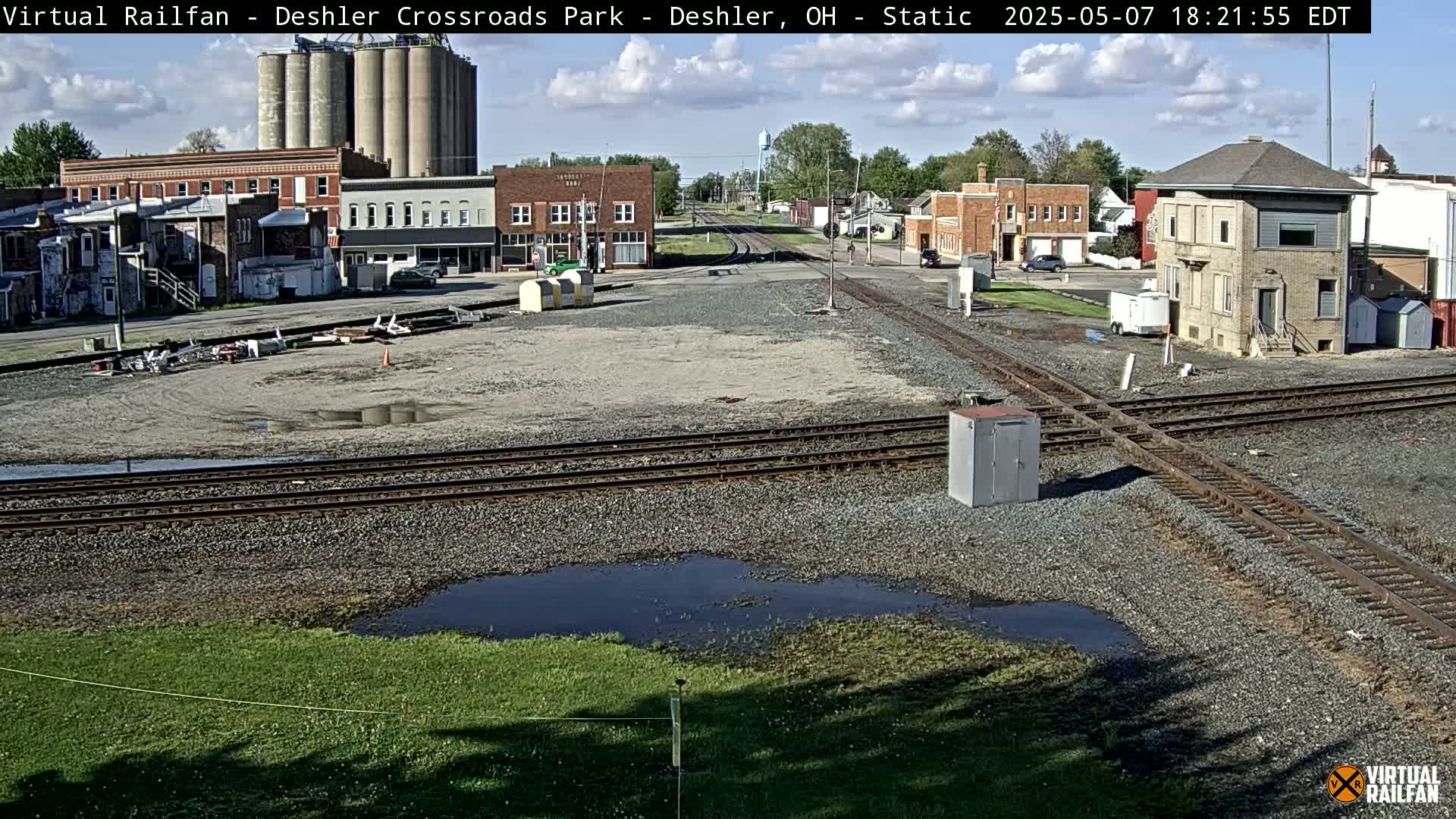 A sunny view of a railroad crossing in a small town, featuring several buildings, grain silos, and a grassy area with puddles of standing water.