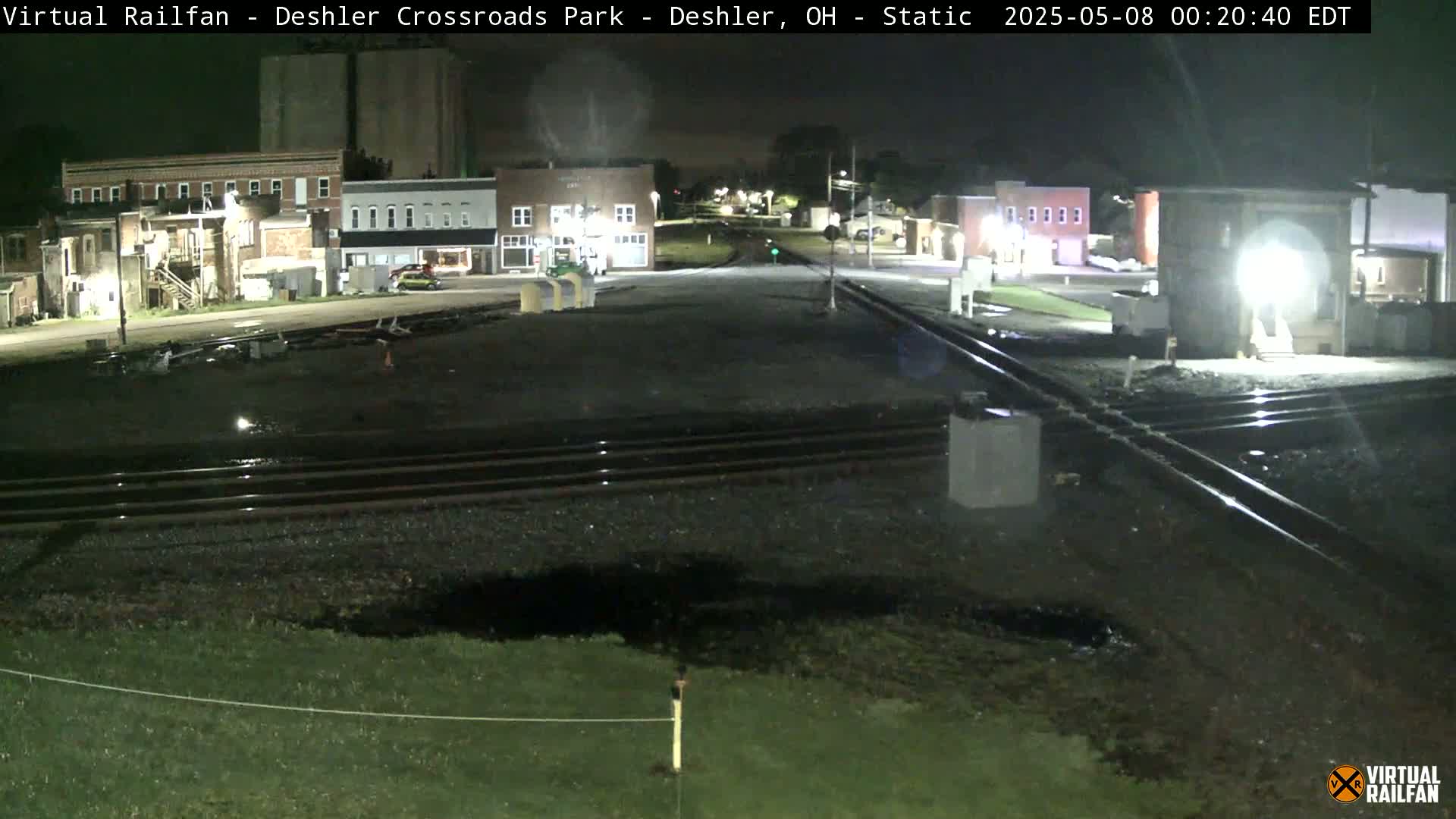 A nighttime view of a railroad crossing near several buildings, with wet ground and train tracks visible.