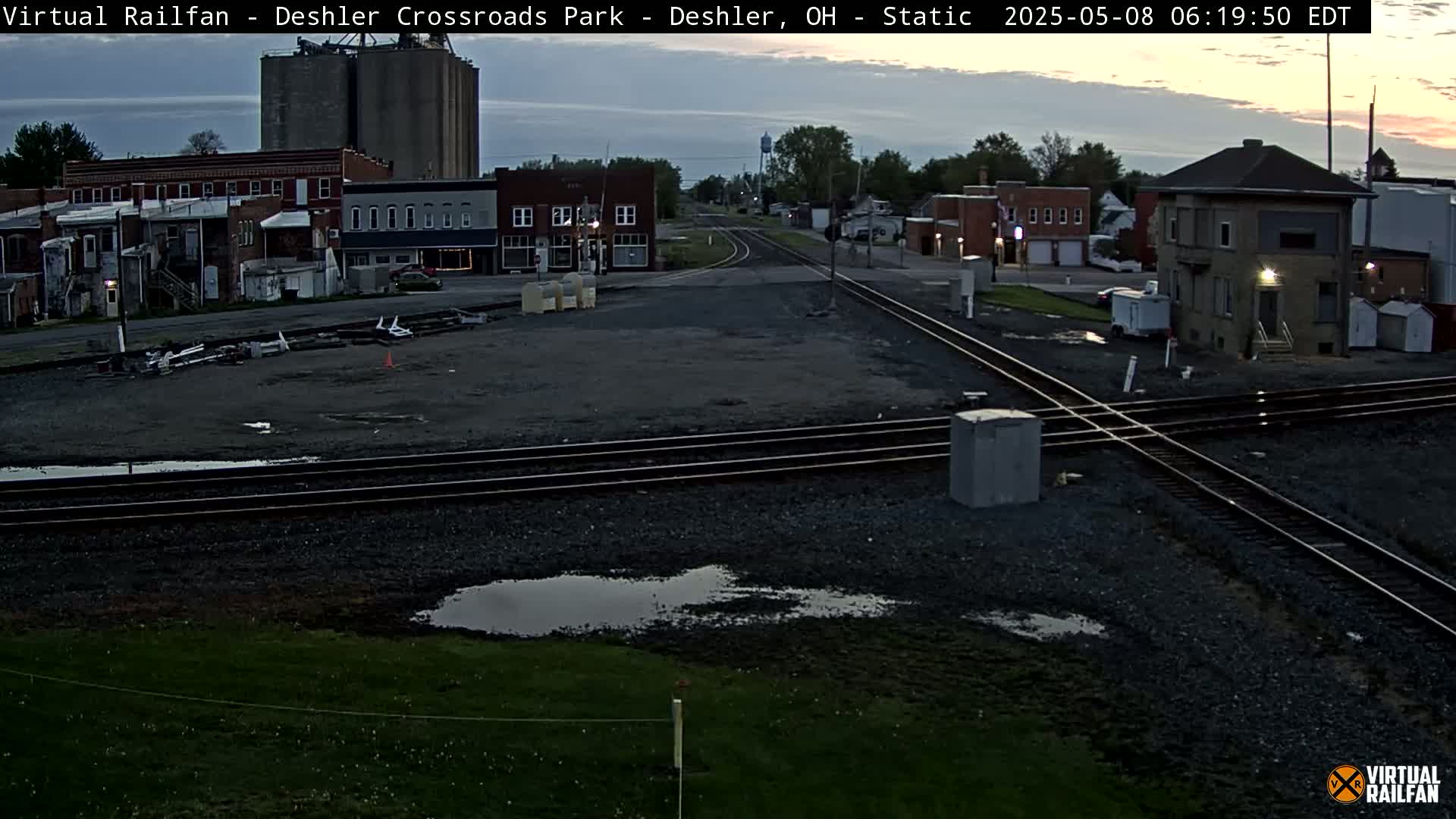 A railroad crossing in a small town at dawn, with puddles on the ground and buildings in the background.