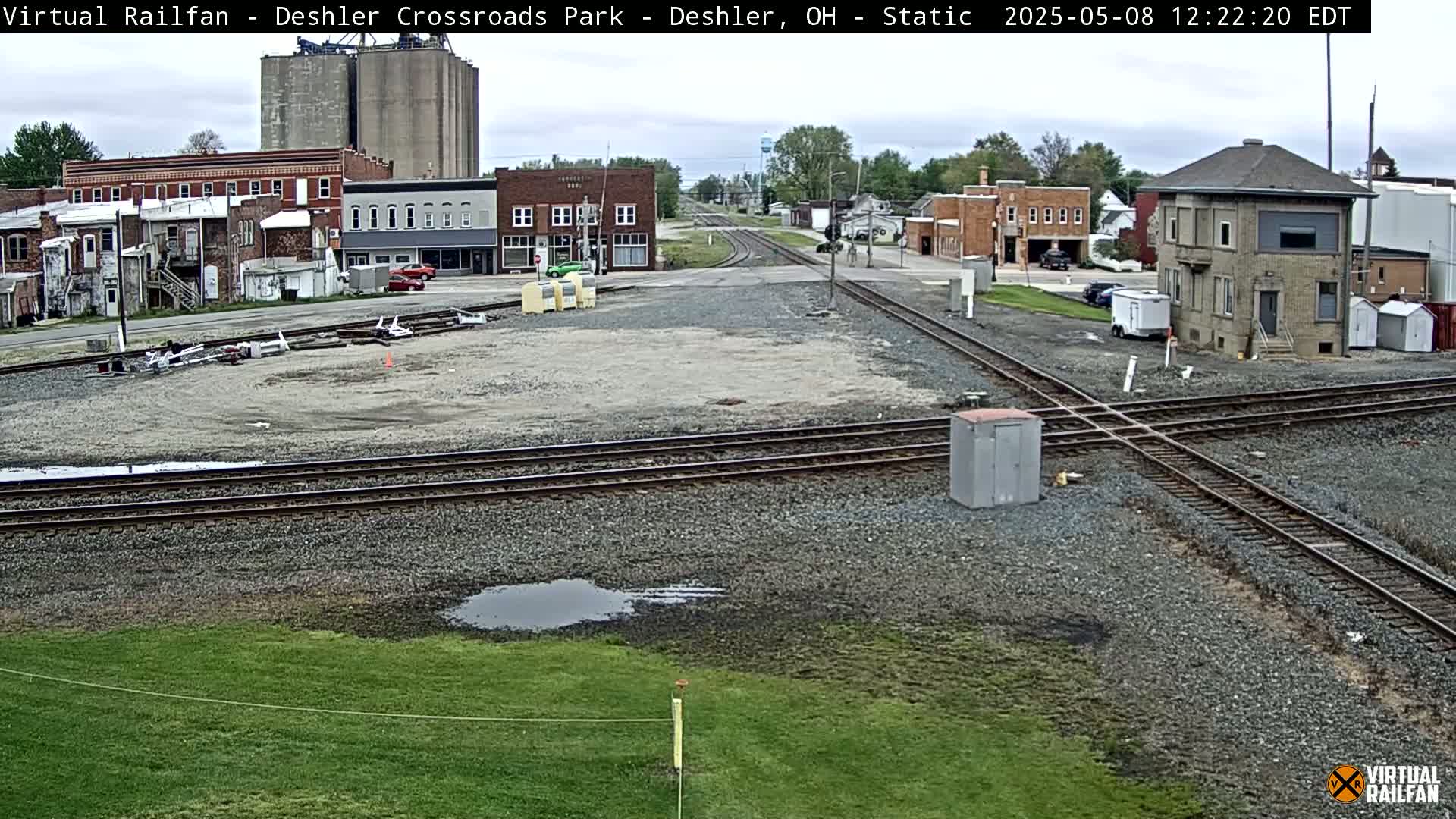 A train crossing in a small town under an overcast sky, with several buildings and a small grass area visible.