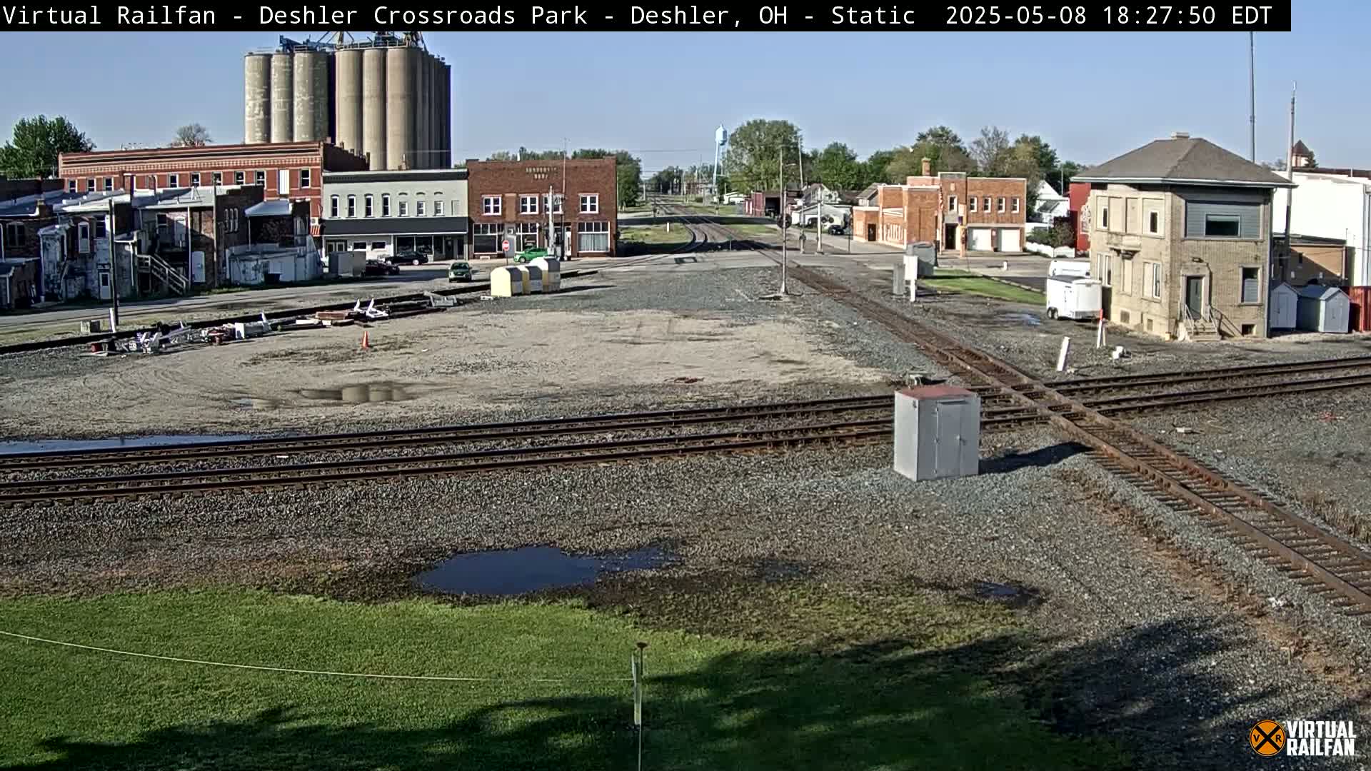 A sunny view of a railroad crossing in a small town, with several buildings, grain silos, and puddles of water visible on the ground.