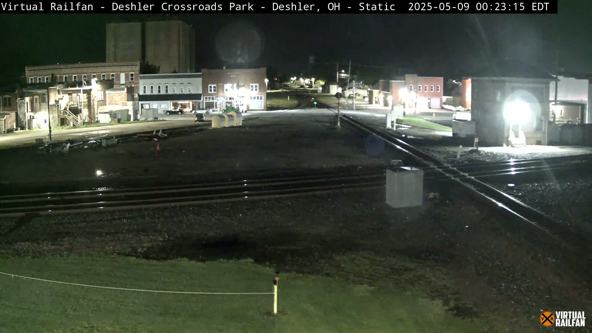 A nighttime view of railroad tracks intersecting a road in front of several buildings under clear skies.