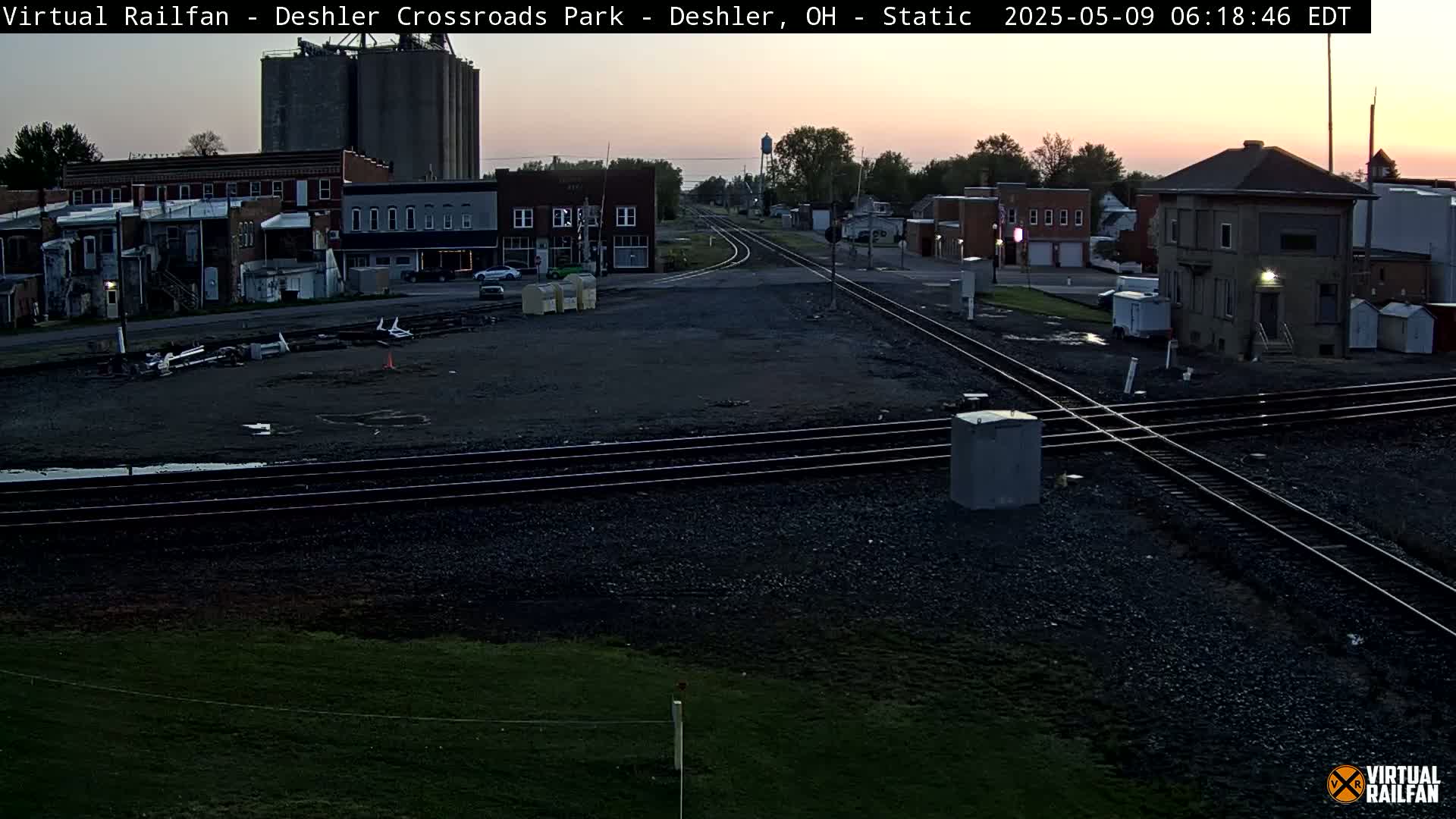 A dusk-lit railroad crossing in a small town, with several buildings and grain silos visible in the background.