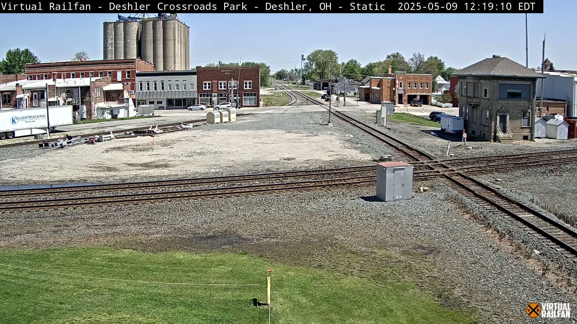 A sunny day shows a railroad crossing in a small town with grain silos, buildings, and a few cars visible.