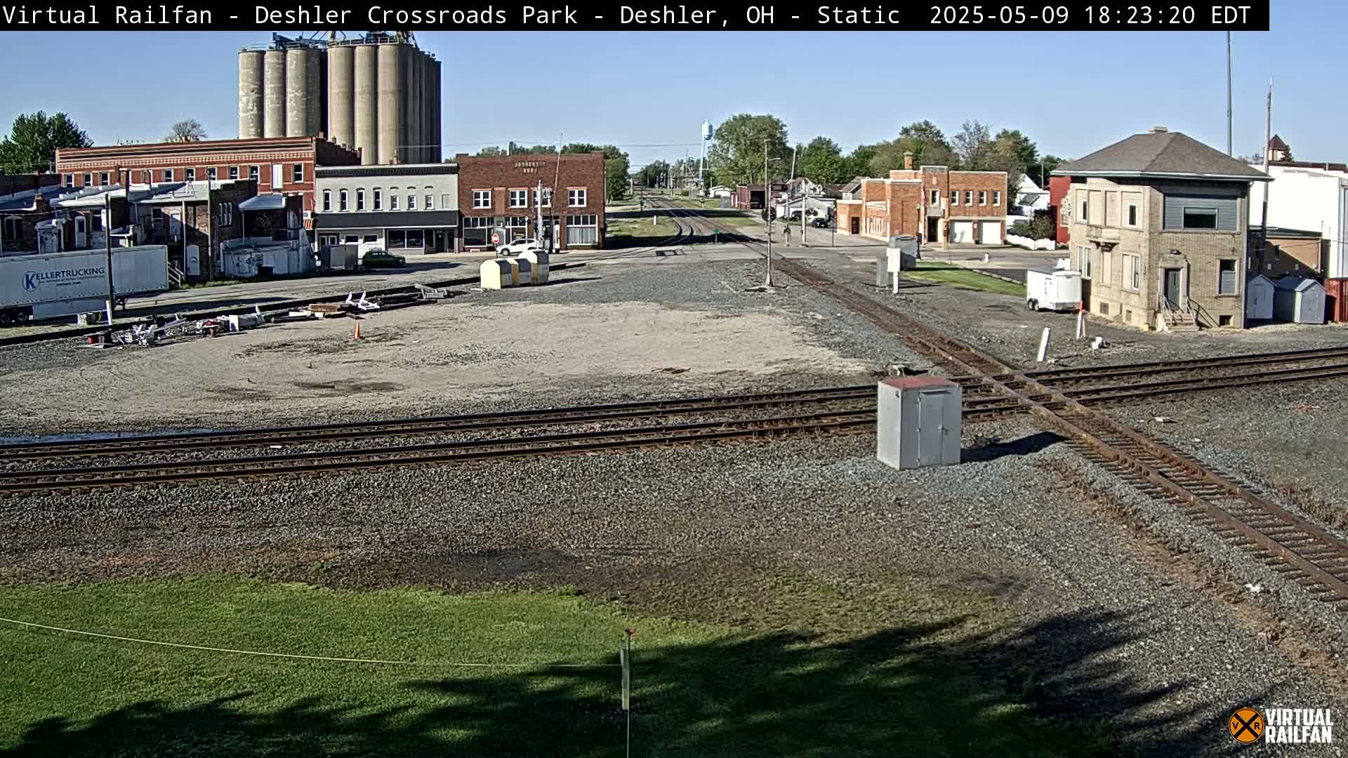 A small town with grain silos, buildings, and a railroad crossing is shown on a sunny day.