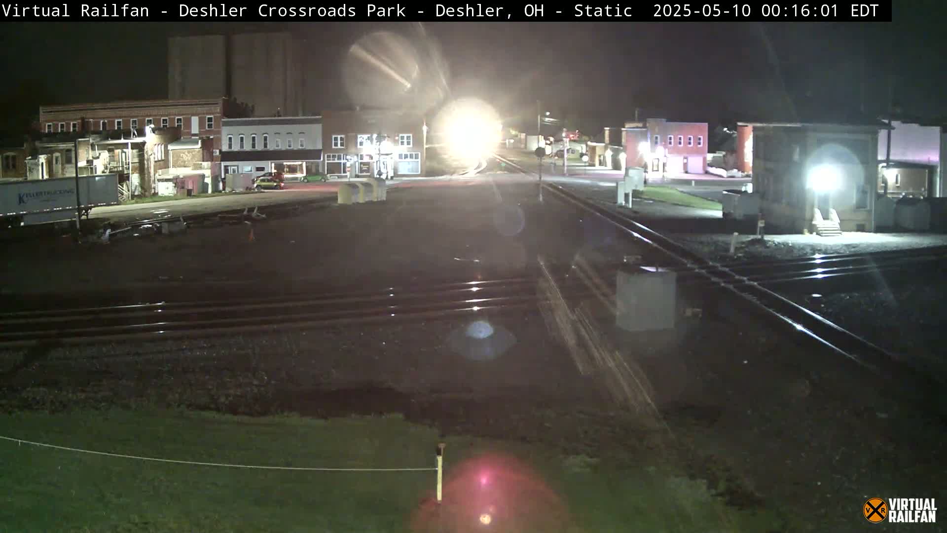 At night, a nighttime view shows train tracks intersecting a road in front of several buildings, with a headlight in the distance.