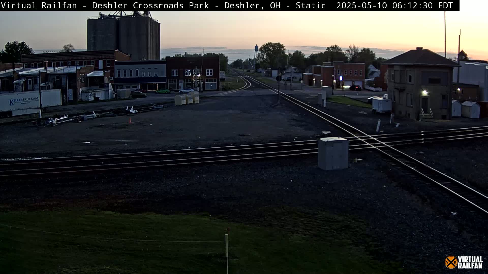 A train crossing in a small town at dawn, with a few buildings and grain silos visible in the background.