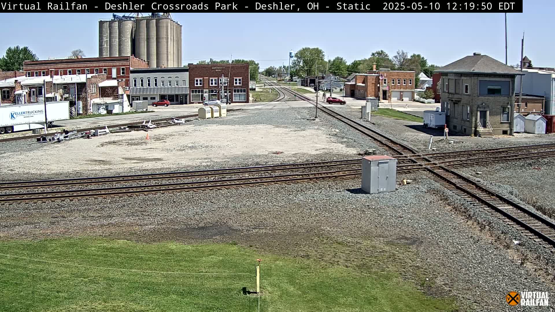 A sunny day reveals a railroad crossing in a small town, with grain elevators, brick buildings, and a rail junction visible.