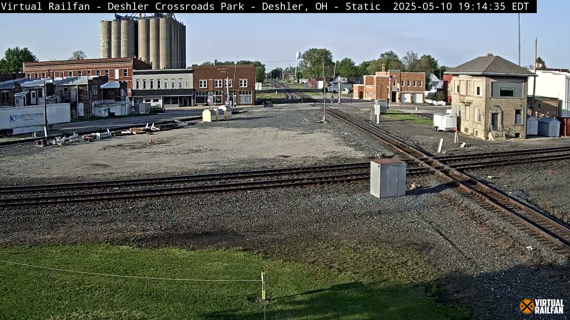 A sunny view of a railroad crossing in a small town, showing several buildings, grain silos, and intersecting train tracks.