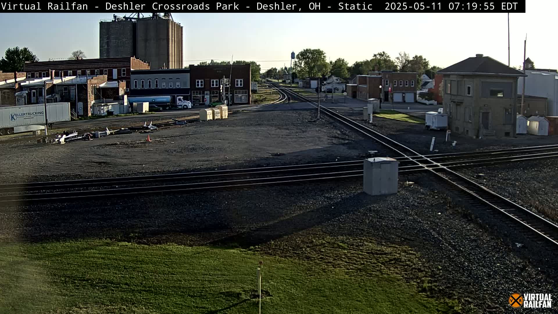 A sunny view of a railroad crossing in a small town, with several buildings and grain silos in the background.