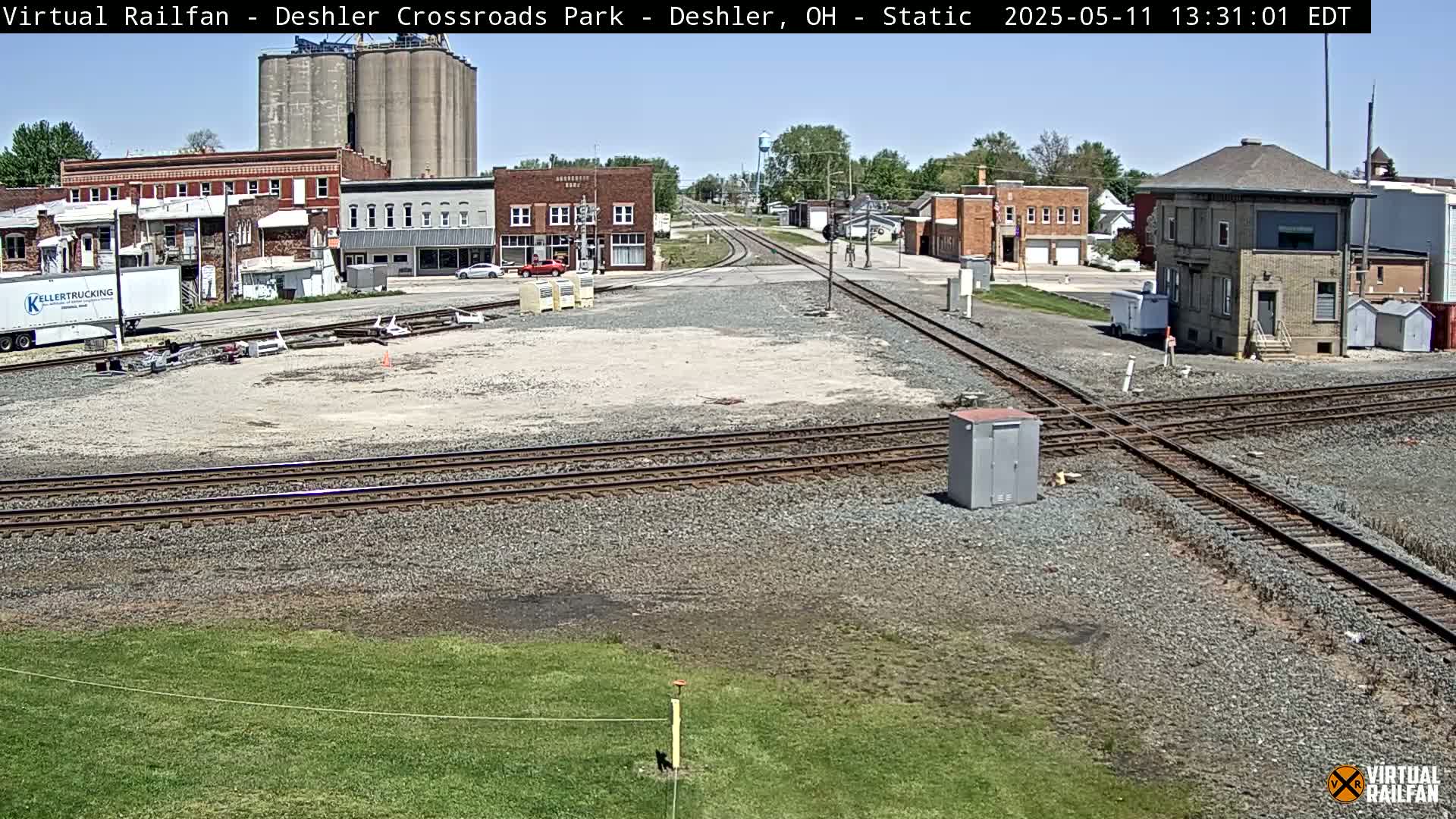 A sunny day reveals a railroad crossing in a small town, with several buildings, grain silos, and a rail yard visible.