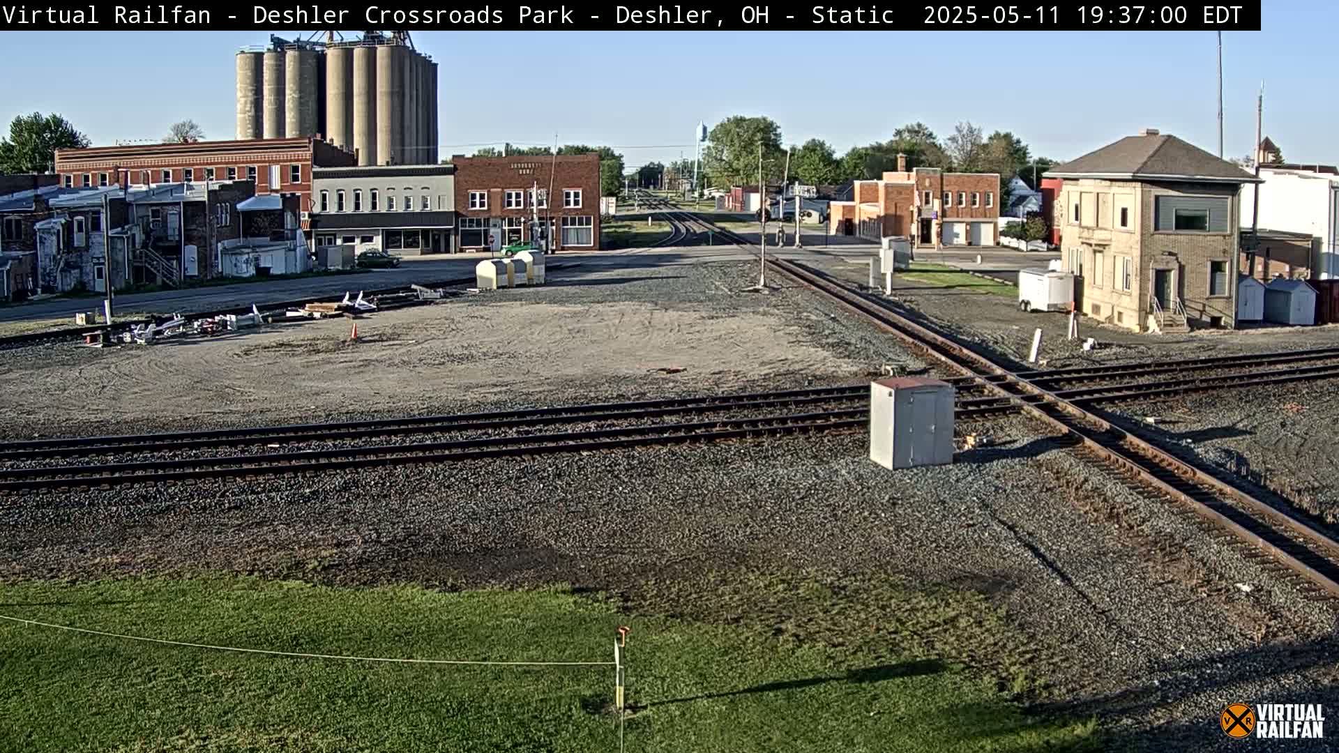 A mostly sunny view of a small town's railroad crossing, showing several buildings, grain silos, and intersecting train tracks.