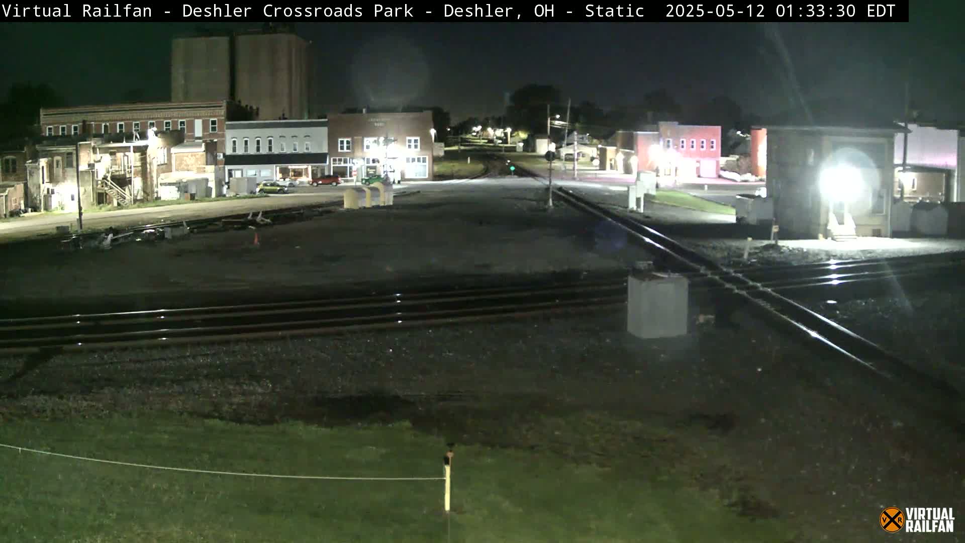 At night, a railroad crossing in a small town features several buildings, railroad tracks, and sparse vegetation.