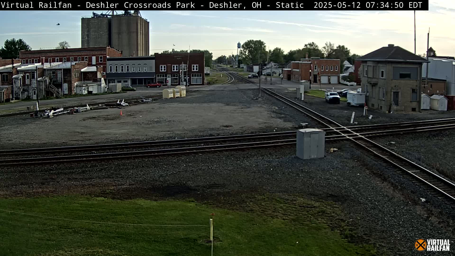 A mostly clear daytime view of a train crossing in a small town, with several buildings, grain silos, and some parked vehicles visible.