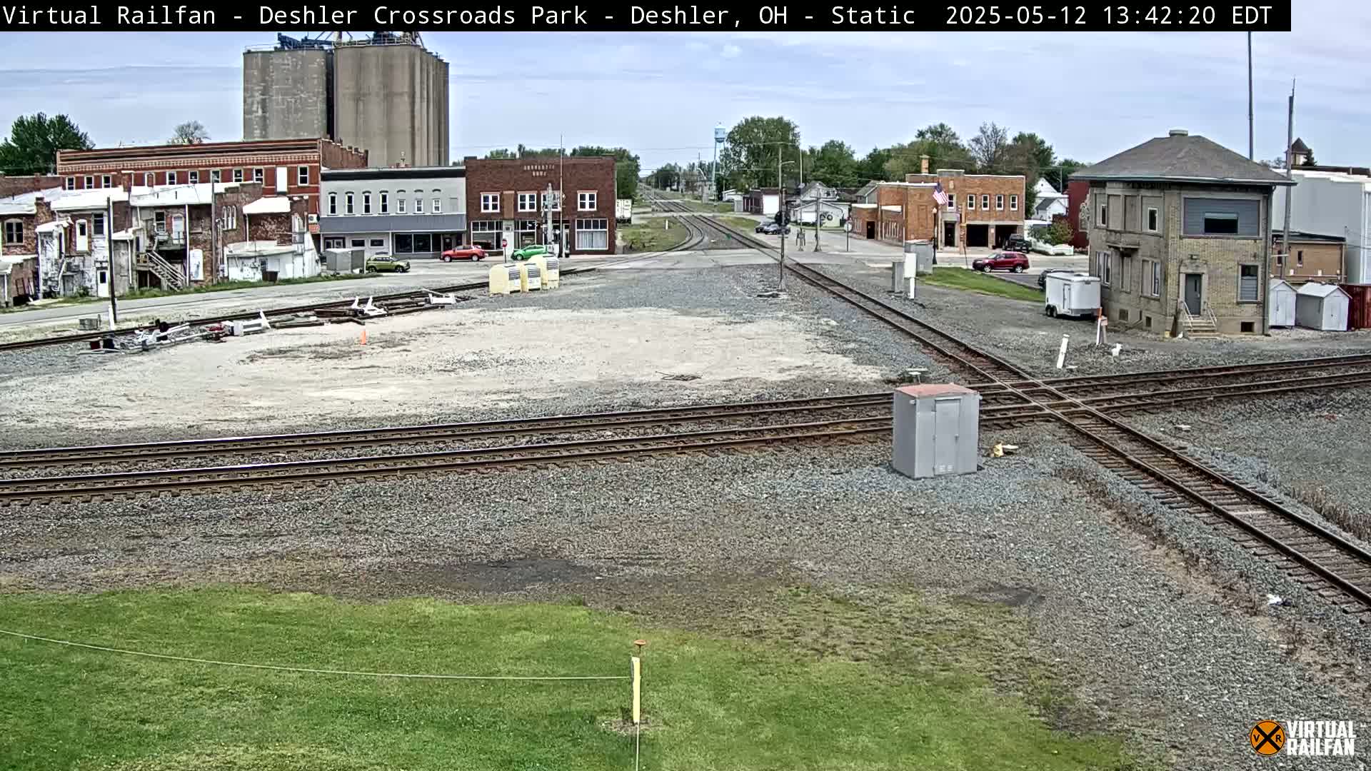 A railroad crossing in a small town on a partly cloudy day, with several buildings and cars visible in the background.