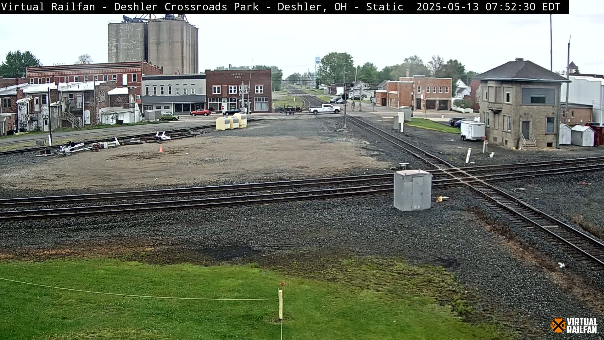 A railroad crossing in a small town on an overcast day, with several buildings and a few vehicles visible.