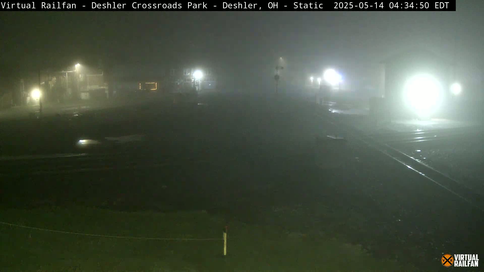 A foggy night scene shows a railroad crossing with dimly lit buildings and train tracks in the distance.