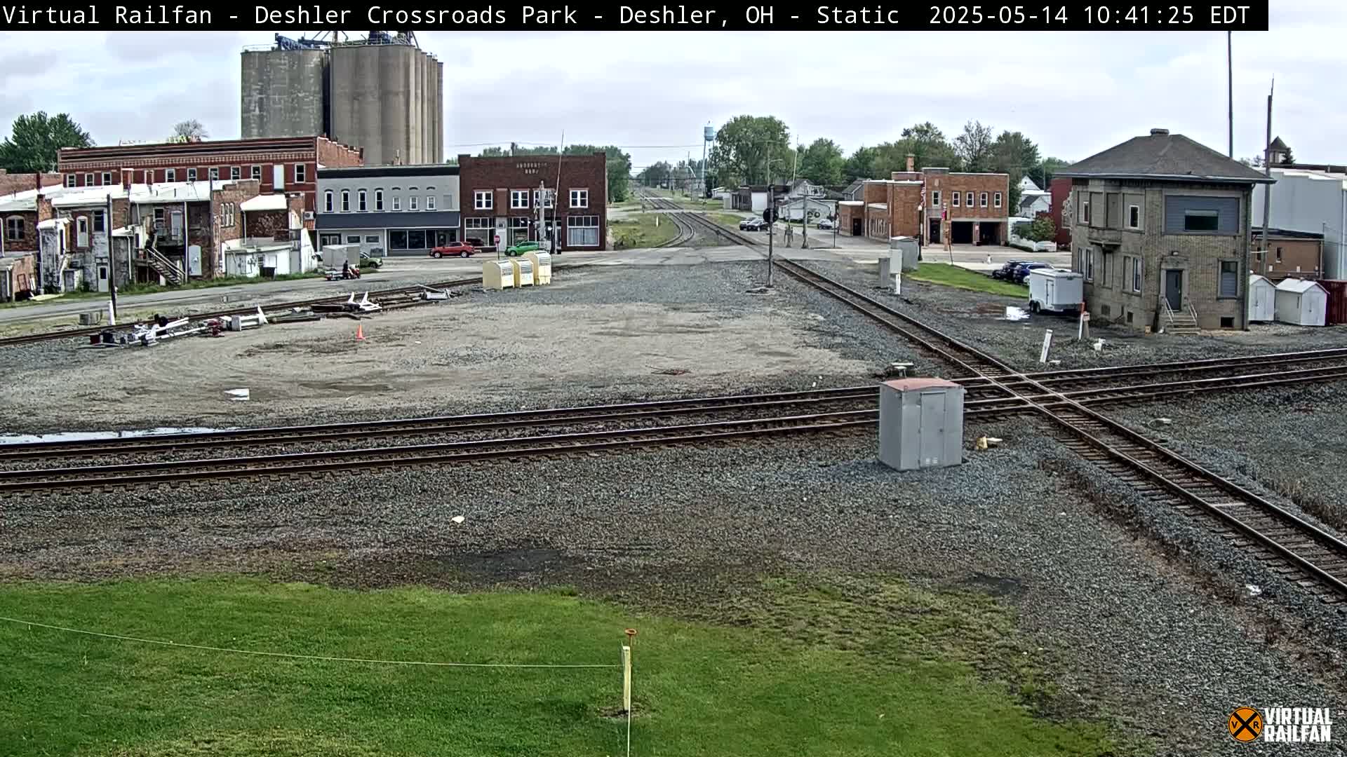 A railroad crossing in a small town on an overcast day, with several buildings and grain silos visible in the background.