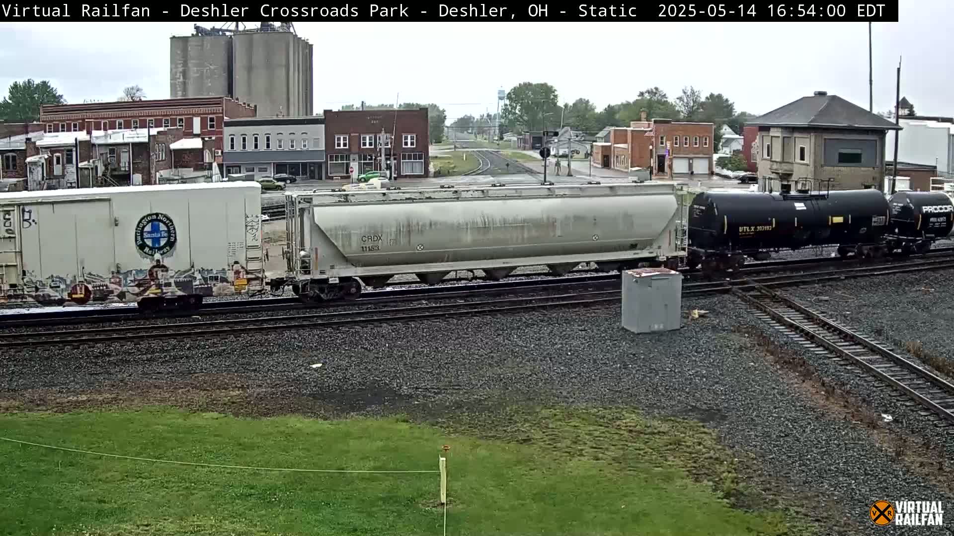 A freight train, consisting of boxcars and tank cars, sits on railway tracks in front of a small town under an overcast sky.