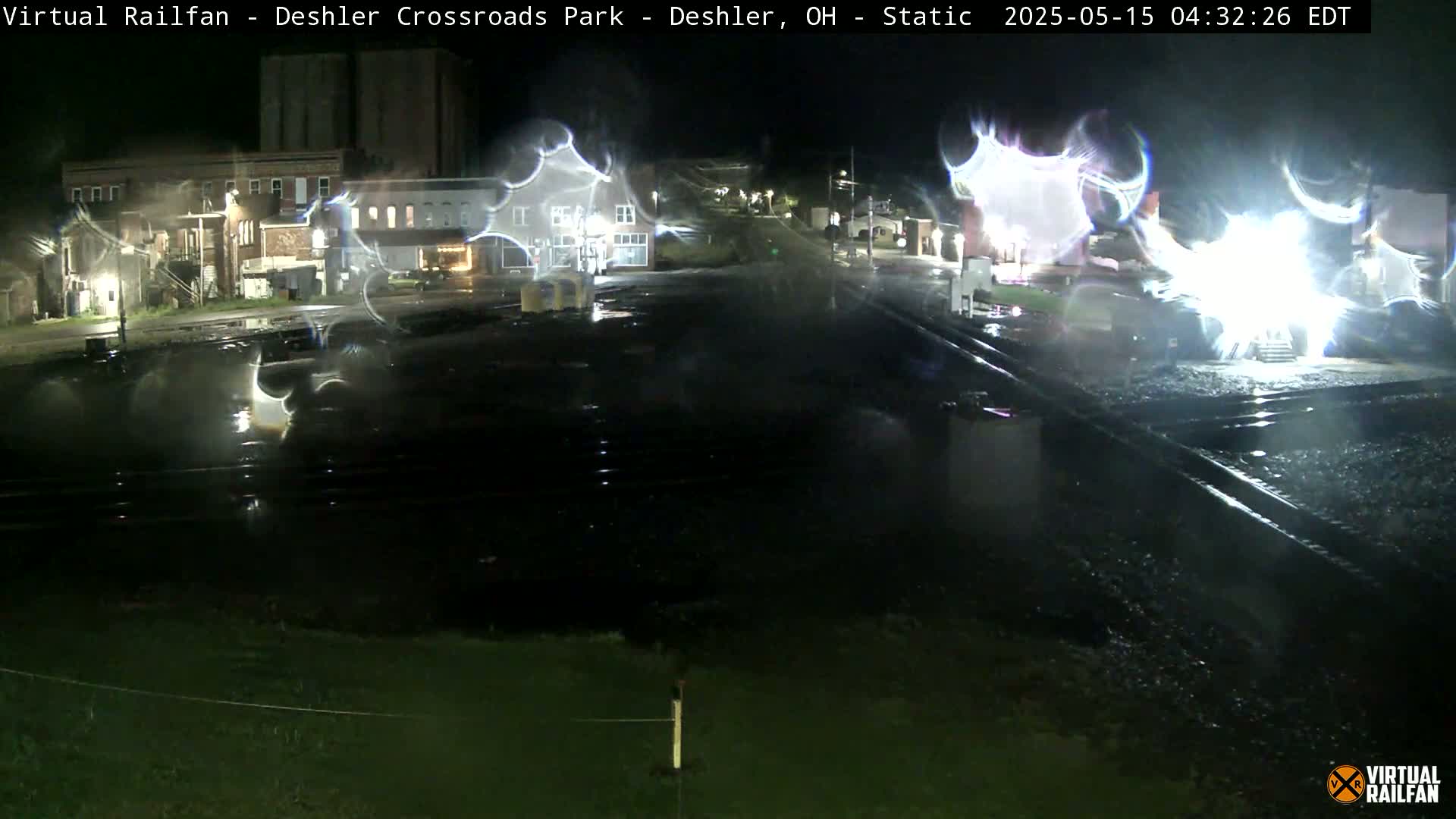 A nighttime view of wet railroad tracks and nearby buildings, illuminated by streetlights and possibly rain.