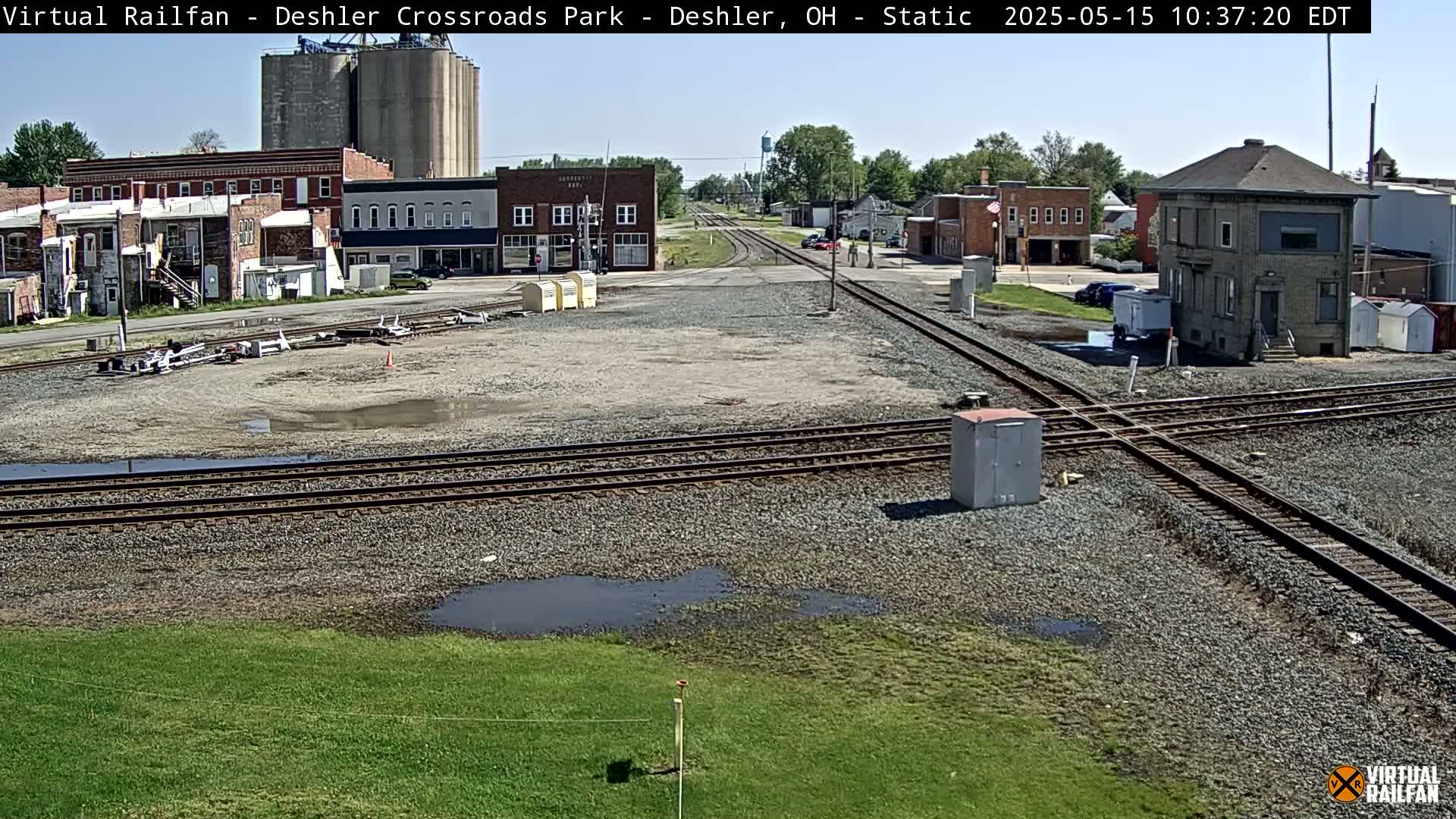 A railroad crossing in a small town on a partly sunny day shows several buildings, grain silos, and a rail yard with puddles of water.
