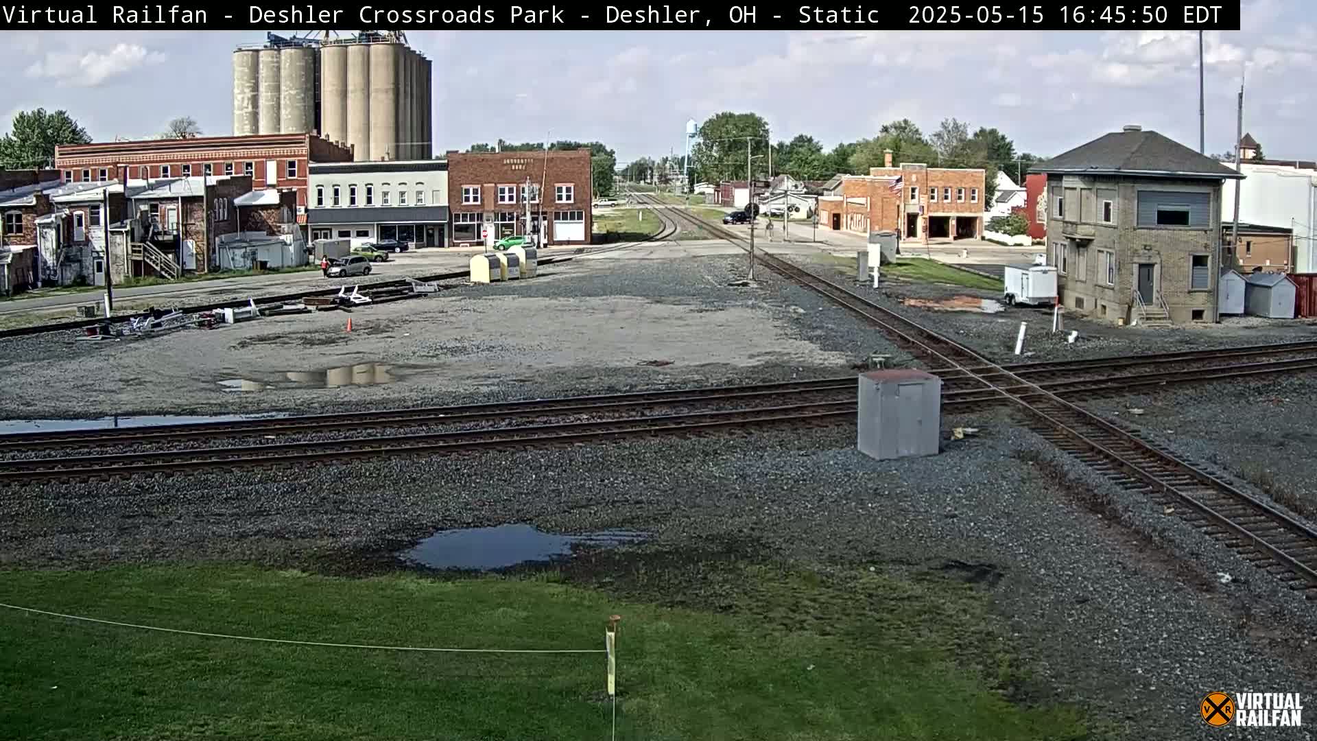 A mostly sunny view of a railroad crossing in a small town, with several buildings, grain silos, and some puddles of water on the ground.