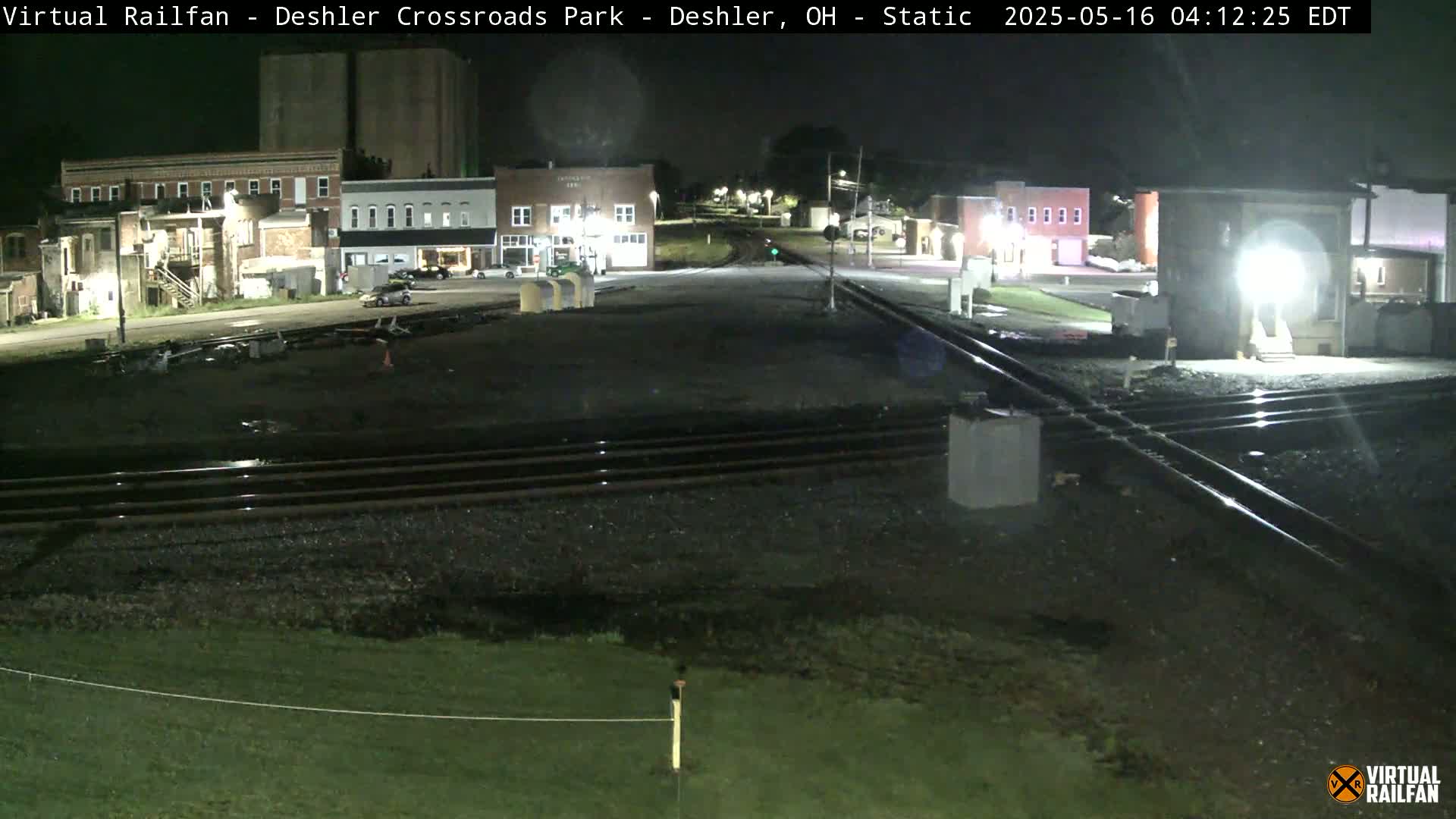 A nighttime view of a railroad crossing in a small town, with several buildings visible under artificial light.