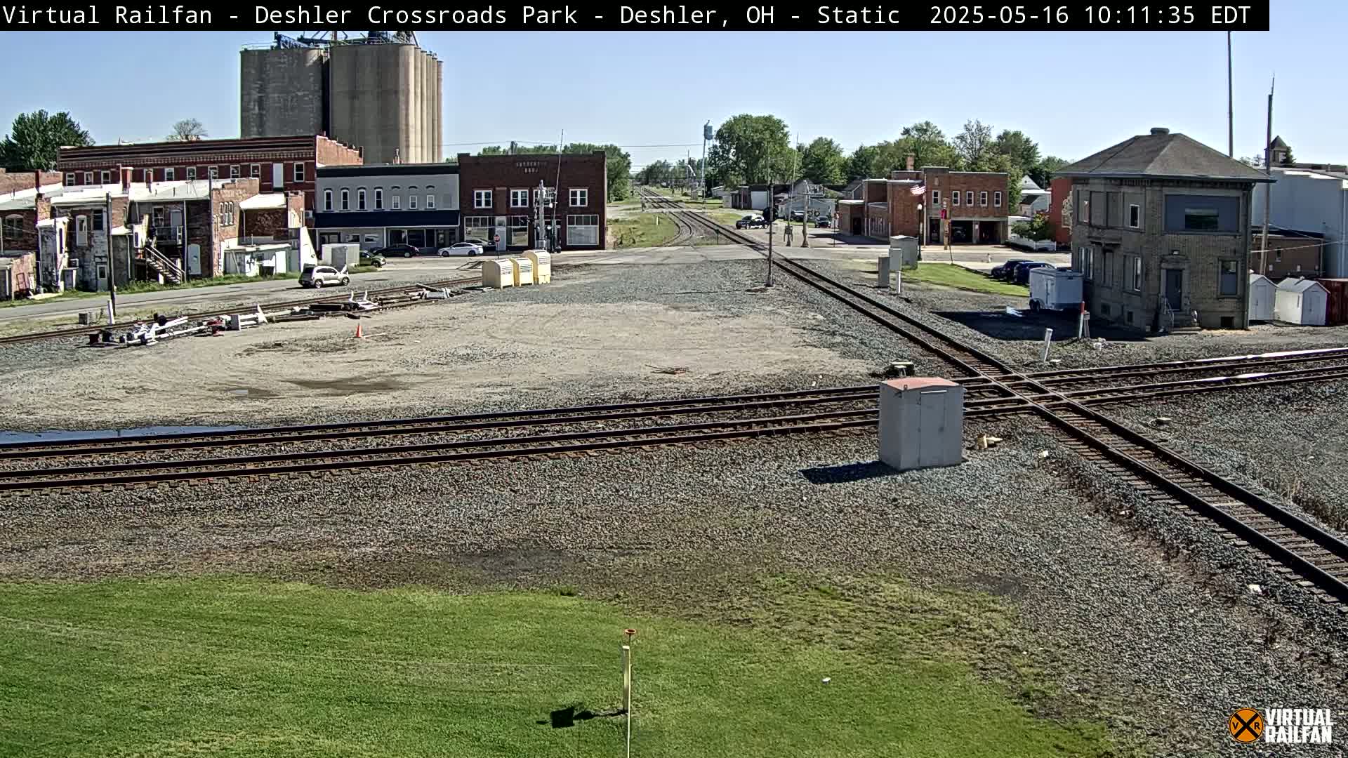 A sunny view of a railroad crossing in a small town, with buildings and grain silos visible in the background.
