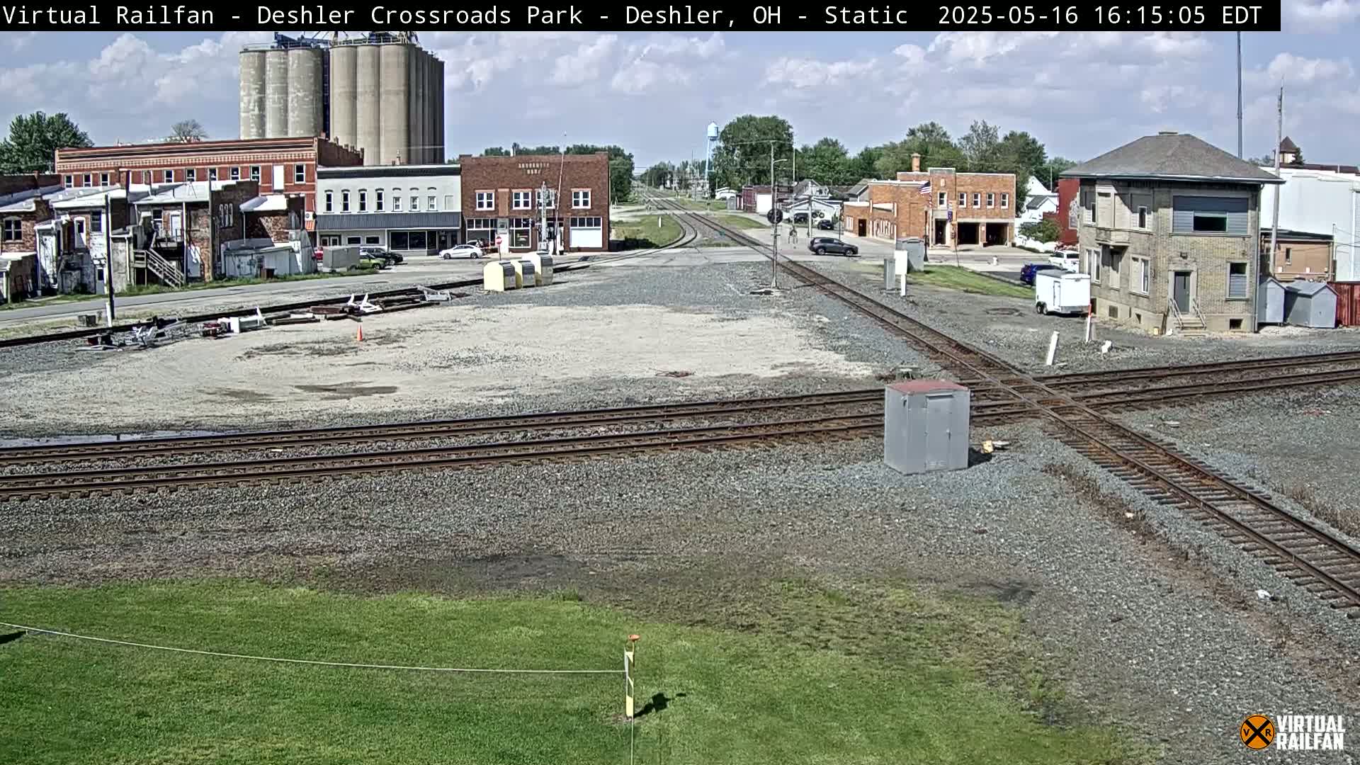A sunny day reveals a train crossing in a small town with several buildings, grain silos, and intersecting railroad tracks.