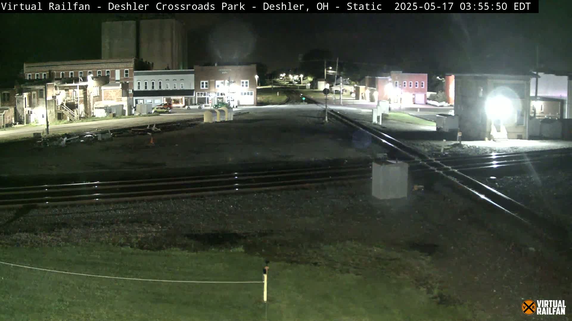 A nighttime view of a railroad crossing in a small town, with several buildings visible under artificial light.