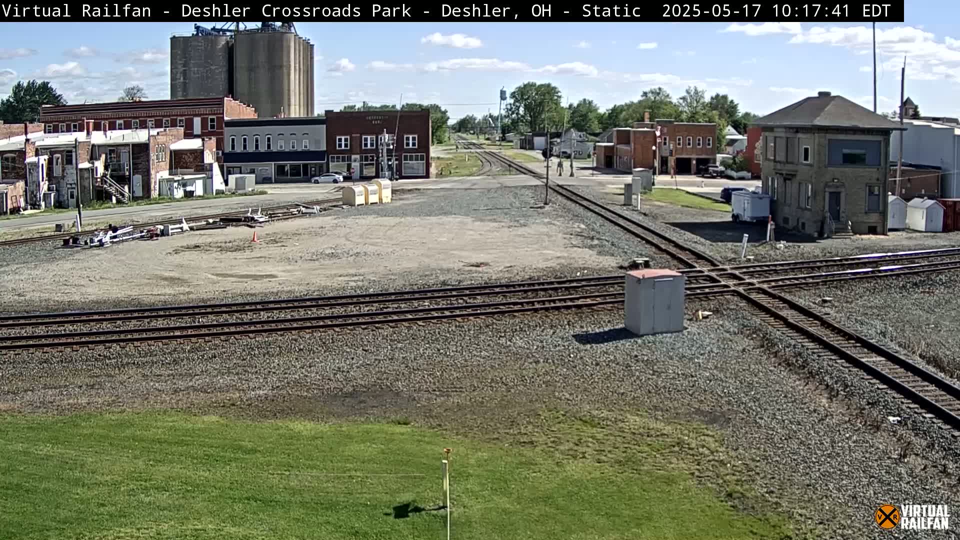 A sunny day shows a railroad crossing in a small town with several brick buildings and grain silos in the background.