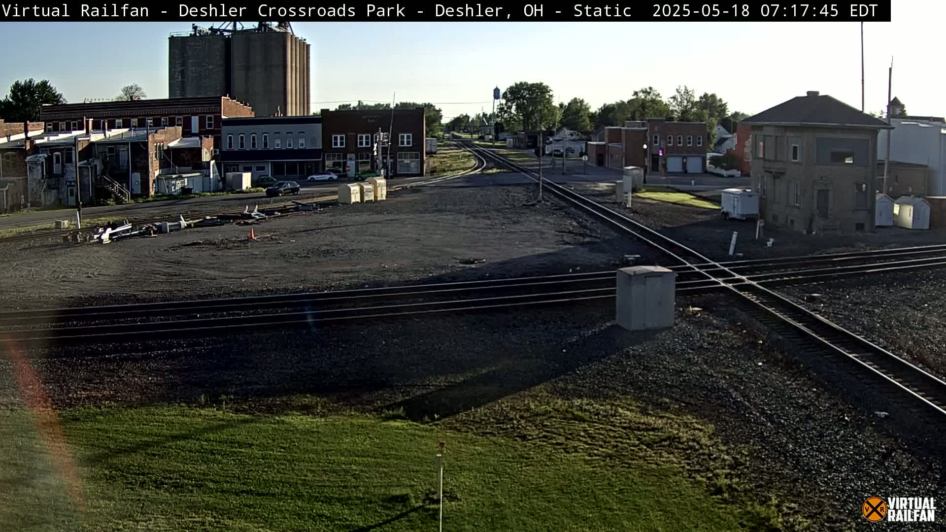 A railroad crossing in a small town on a sunny morning, with several buildings and grain silos visible in the background.
